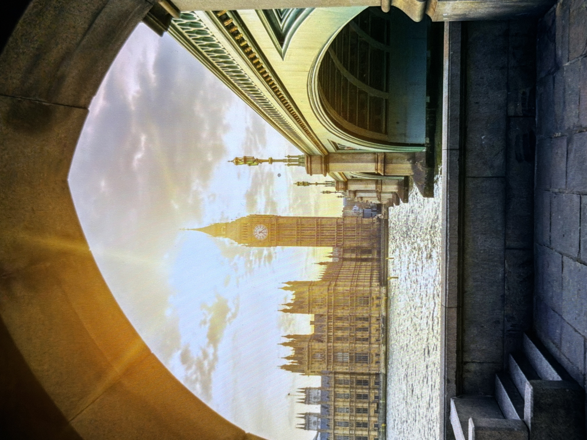 The scene depicts a notable architectural ensemble situated by a wide river in the city of Maidstone, United Kingdom. The foreground is defined by a heavy stone archway, framing the view. In the mid-ground, a large, ornate Gothic Revival building, characterized by numerous spires, turrets, and intricate detailing, stands on the far bank of the river. Adjacent to it on the right, a very tall, square clock tower with a prominent clock face rises into the sky, crowned with a spire. Spanning the river on the right is an elaborately designed multi-arched bridge, featuring decorative green and white railings and period streetlamps with golden tops. The river surface displays reflections and ripples from the overhead light. The immediate lower-left foreground shows a stone embankment with a short flight of three steps, while the lower-right foreground consists of paved stone slabs. The sky above is partly cloudy, illuminated by a strong, radiant light source, possibly the sun, positioned behind the clock tower, creating visible light rays and a bright glare. No individuals are present in the visible portion of the image.