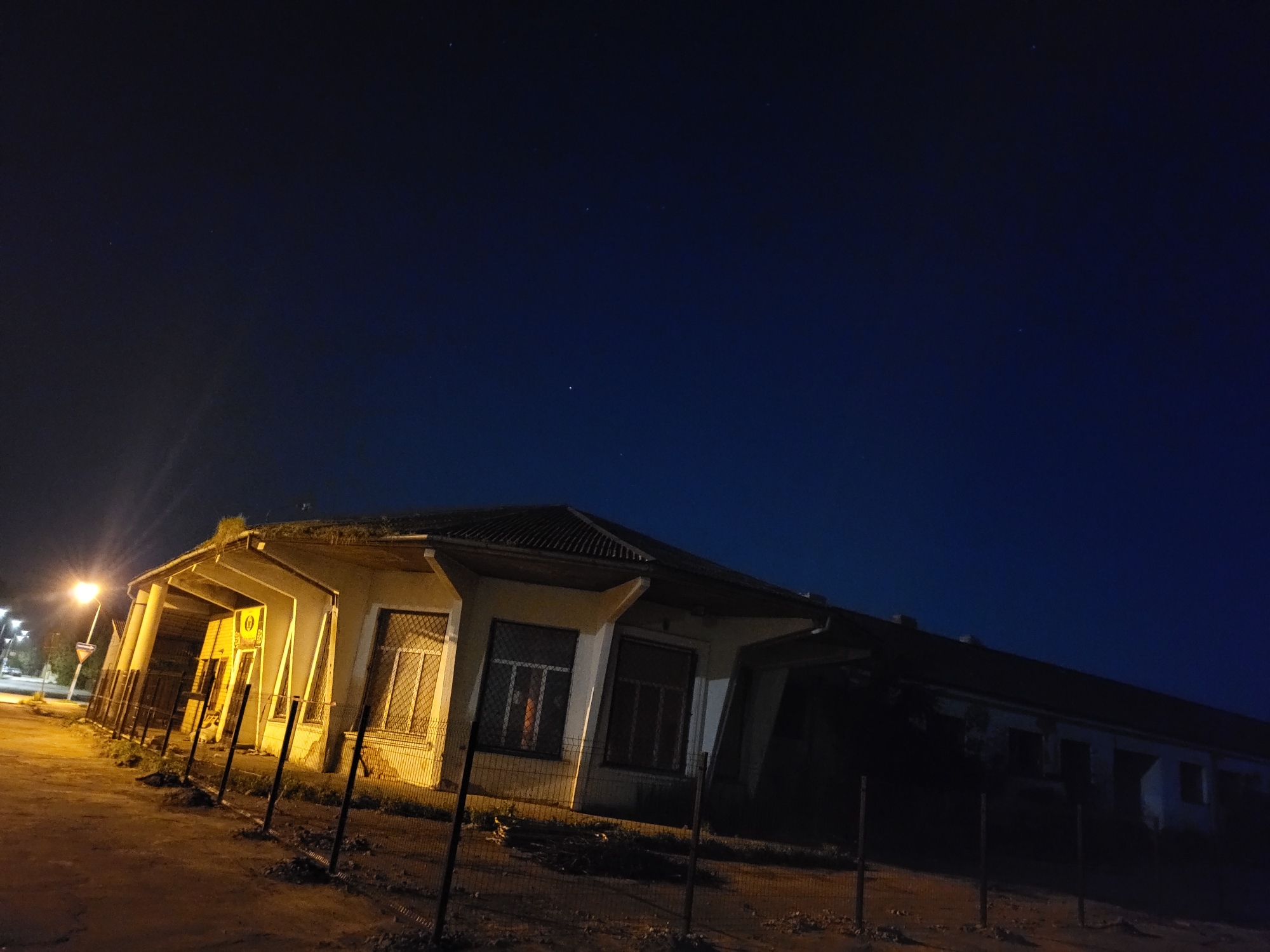 An outdoor, eye-level, wide shot shows an abandoned, single-story building at night in Jelgava, Latvia. The building is illuminated by a streetlamp and lights from within the structure. The building has a dark, slanted roof and several windows with metal grates. A chain-link fence runs along the front of the property. The sky above is a deep blue and shows several stars. The ground in front of the building is dirt and gravel.
