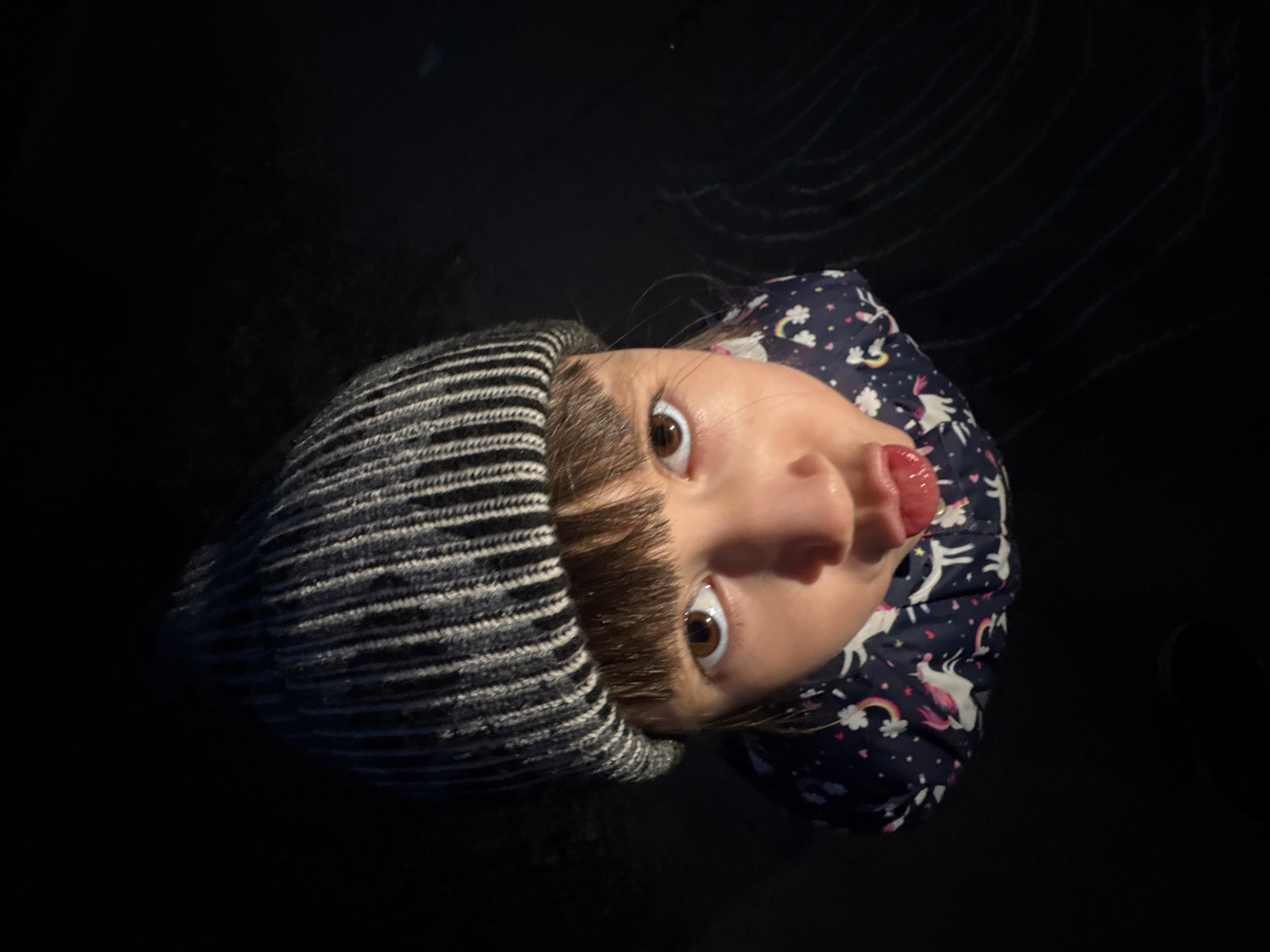 A young child is captured in a close-up, upward-angled shot against a predominantly dark background. The child, with light brown hair peeking from under a striped grey and white beanie, is looking directly at the camera with wide, expressive brown eyes. A playful emotion is conveyed by the child sticking out their tongue. They are wearing a dark blue jacket adorned with white unicorn and rainbow patterns.
The background is almost entirely black, with subtle hints of texture and faint, light-colored lines, particularly visible in the upper right portion of the image. These lines resemble chalk drawings on a dark, paved surface, suggesting an outdoor setting under low light conditions, possibly in Chaikovsky, Russia. The perspective implies the camera is held above the child, looking down. The child's attire, including the beanie and jacket, suggests cool weather. There is no visible text in the image.