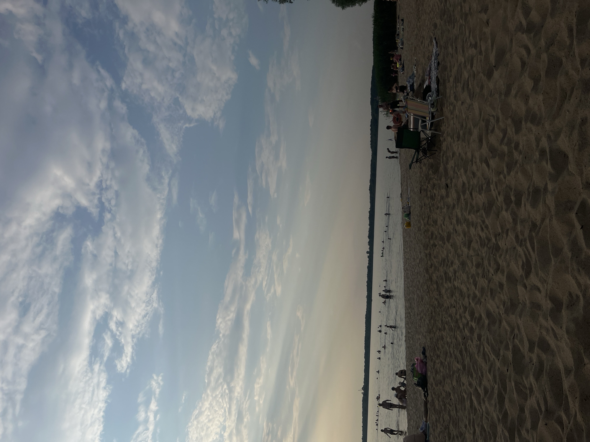 A wide shot of a sandy beach on a cloudy day. Numerous people are in the water, some swimming and others wading. On the shore, people are relaxing on towels, and there are a couple of beach chairs. The water extends to a distant tree line under a sky with scattered clouds and sunbeams.