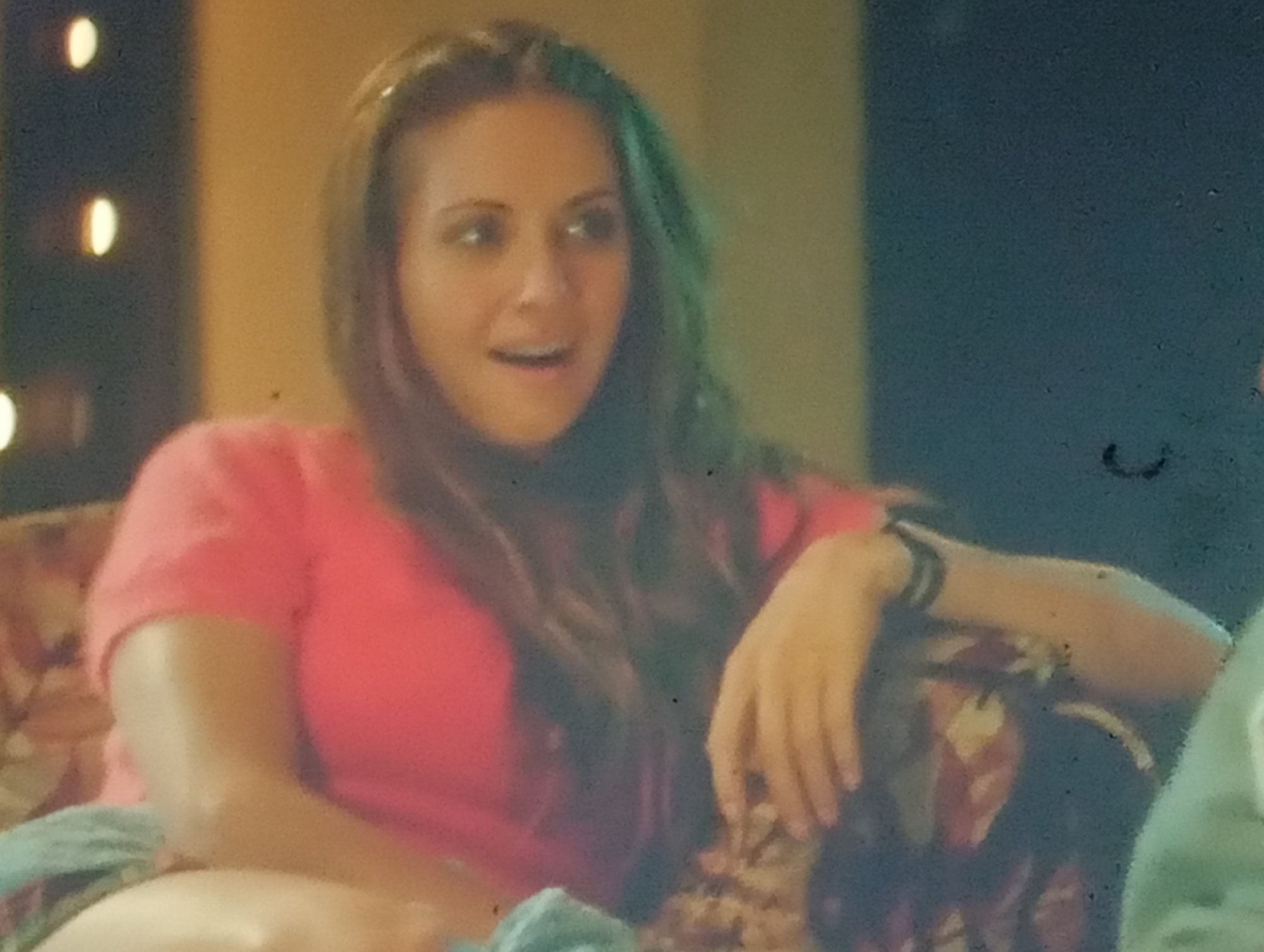 A woman with long, dark, wavy hair, some strands of which appear to have a green tint, is seated on a patterned sofa. She is wearing a bright pink t-shirt. Her mouth is open, as if she is speaking or reacting to something. Her left arm is resting on the back of the sofa, and she is wearing a black bracelet on her wrist. The background is dark with some out-of-focus lights that are oval in shape. This scene does not provide any information that would allow for the determination of its location in Sofiivska Borshchahivka, Ukraine.