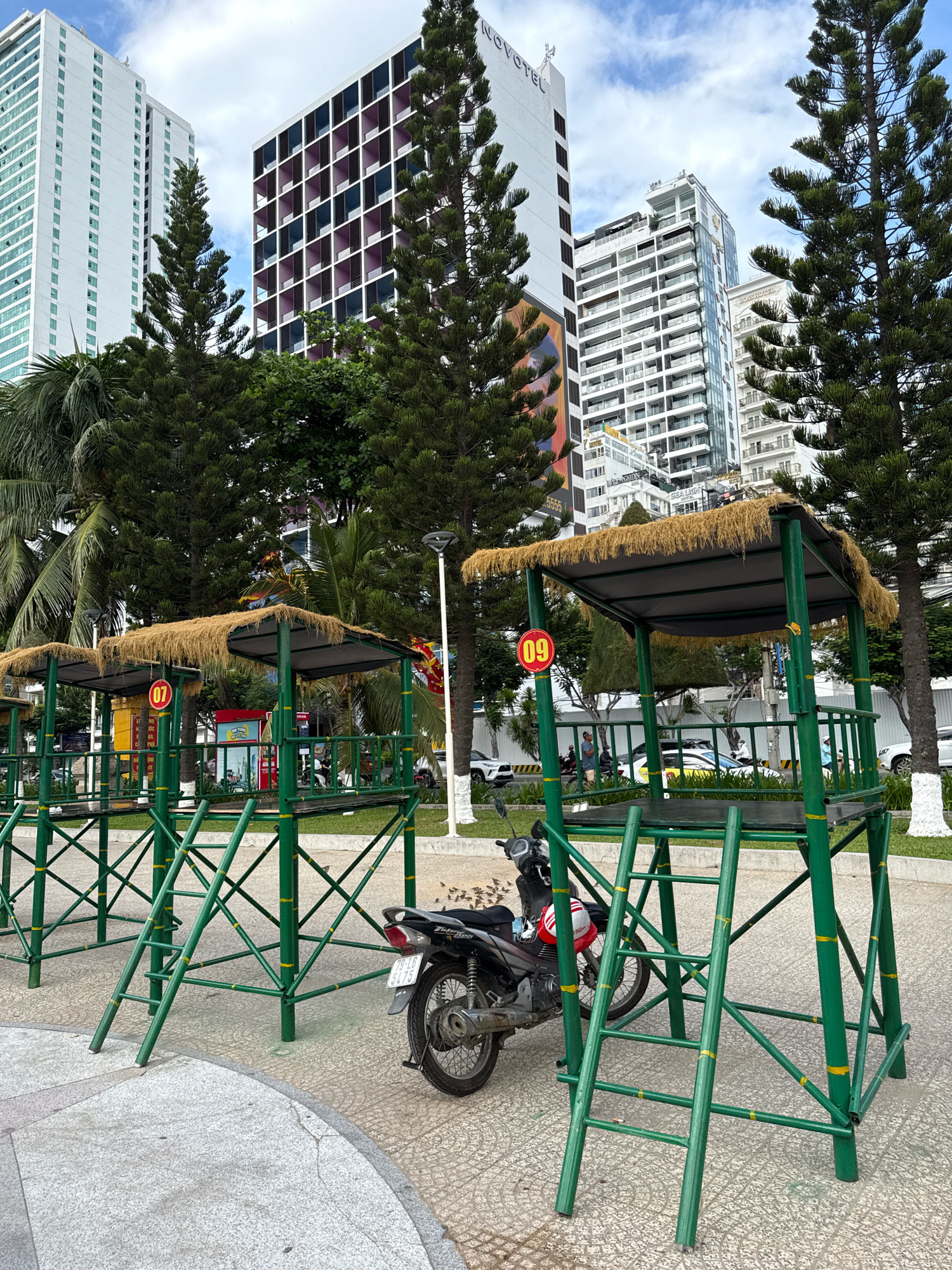 The image depicts a street scene in Nha Trang, Vietnam, featuring a row of elevated, green-painted bamboo-like structures with thatched roofs, likely serving as shelters or observation posts. A black motorcycle is parked in front of one of these structures. In the background, several modern high-rise buildings dominate the skyline under a cloudy sky, with lush green trees and palm trees interspersed. The ground is paved with patterned tiles and some cobblestones. The scene appears to be a public area, possibly near a beach or promenade, given the tropical vegetation and the presence of several buildings. The structures have numbered signs, with '07' and '09' visible on two of them. There are a few individuals visible in the middle ground, and vehicles on a road behind the structures, suggesting an active urban environment.