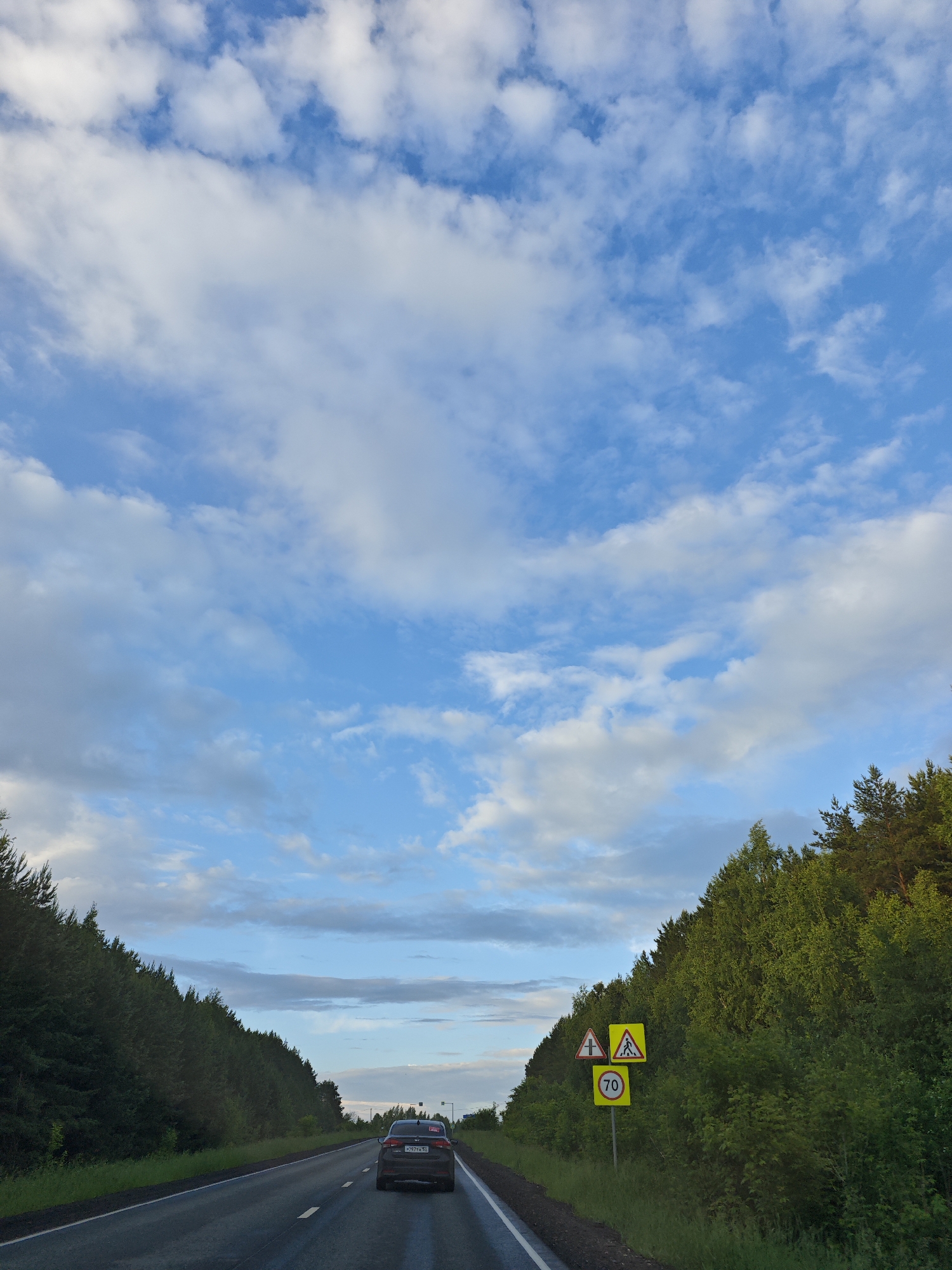 The image captures a view from a vehicle traveling on a paved road through a forested area. The road curves to the right, with a dense line of green trees lining the left side of the frame. On the right side of the road, there is a cluster of road signs. The closest signs are yellow and triangular, depicting a pedestrian crossing and an intersection with a pedestrian symbol. Below these, a circular yellow sign with a red border displays the number "70", indicating a speed limit. Further down the road, another car with a license plate visible is driving ahead. The sky above is blue with scattered white clouds. The scene appears to be in a rural or suburban setting, given the surrounding trees and the absence of urban structures, potentially the area of Новая Казмаска, Russia, based on the provided context.