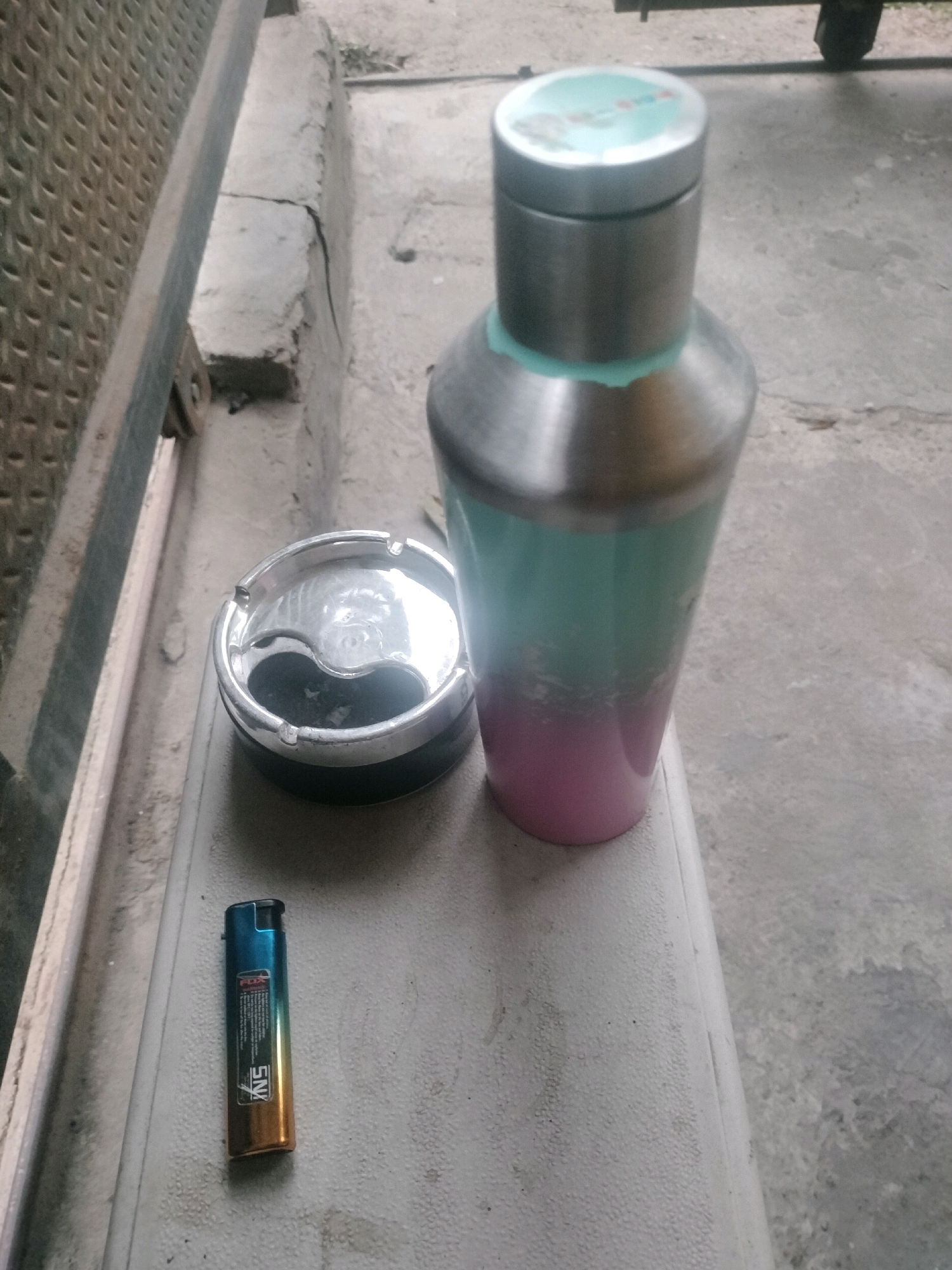 A silver and teal colored metal shaker bottle with a pink gradient, an ashtray, and a blue and gold lighter are placed on a gray, textured surface, possibly a bench or table. The background consists of a concrete area and a metallic structure. The scene appears to be outdoors.