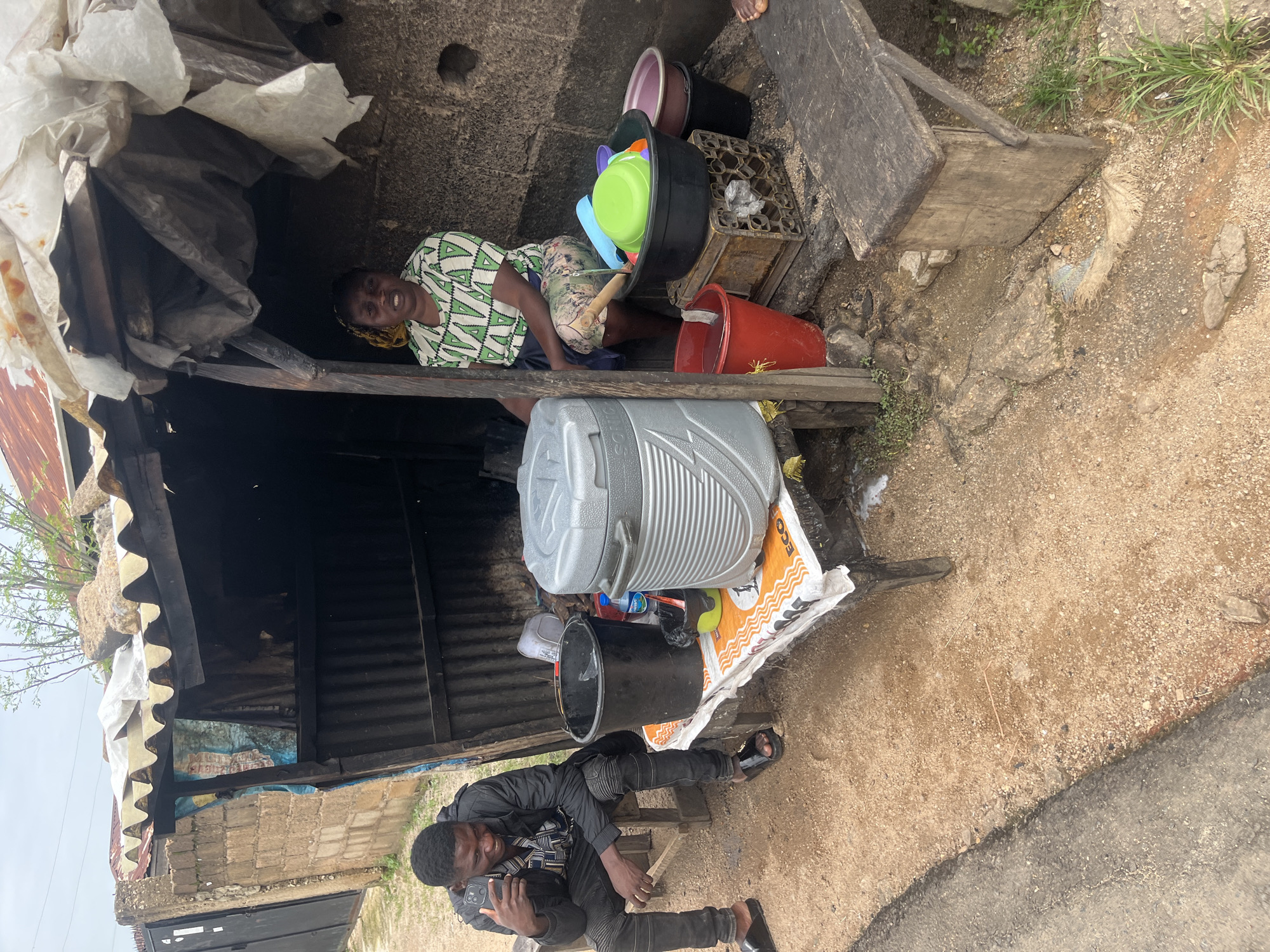 A roadside stall in Jos, Nigeria, displays a man sitting on a wooden bench while holding a smartphone. He wears a black jacket over a patterned shirt and black pants, with black slides on his feet. The stall itself is a rustic structure made of corrugated metal and wood, covered with a white tarp. Inside, a woman in a green and white patterned dress sits, appearing to be the proprietor. In front of the stall, various items are arranged on a table covered with a printed material. These include a large grey cooler, a black bucket, a red bucket with water, and several colorful plastic bowls and basins. The ground is a mix of dirt and gravel. The background shows a stone wall and a partial view of a building with a corrugated metal roof. The overall scene depicts a typical street-side vending setup in an urban environment.