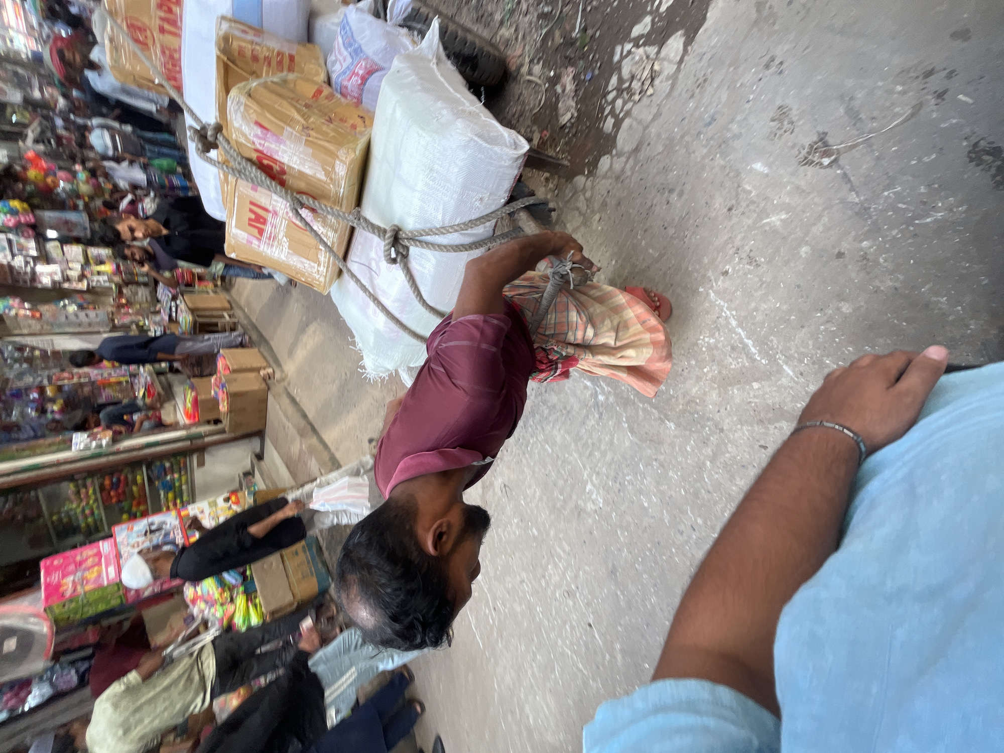 In Dhaka, Bangladesh, a man pulls a cart loaded with packages and bags on a city street. The man wears a maroon shirt and a patterned wrap around his waist. Ropes secure the load on the cart. In the background, shops display various goods. People mill around the shops. The photographer is positioned close to the scene, capturing the man pulling the cart. The street appears to be crowded with pedestrians and vendors.