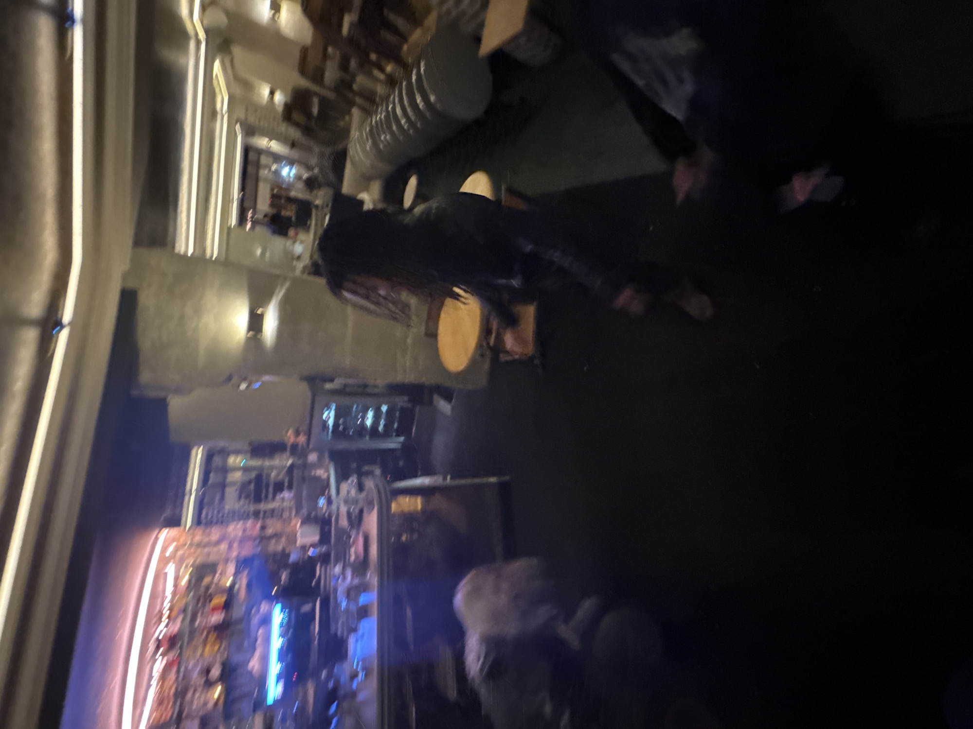 The image depicts a dimly lit interior, seemingly a bar or restaurant. The scene appears to be inside a building with exposed ductwork and various light sources. There is an individual standing and leaning forward on a bar-like structure. Towards the left, there is an illuminated bar area with a variety of bottles and fixtures. Other people can be seen in the background. The overall impression is a casual, urban environment. It's difficult to definitively ascertain the location to Paris, France without further information or distinguishing architectural or cultural details.