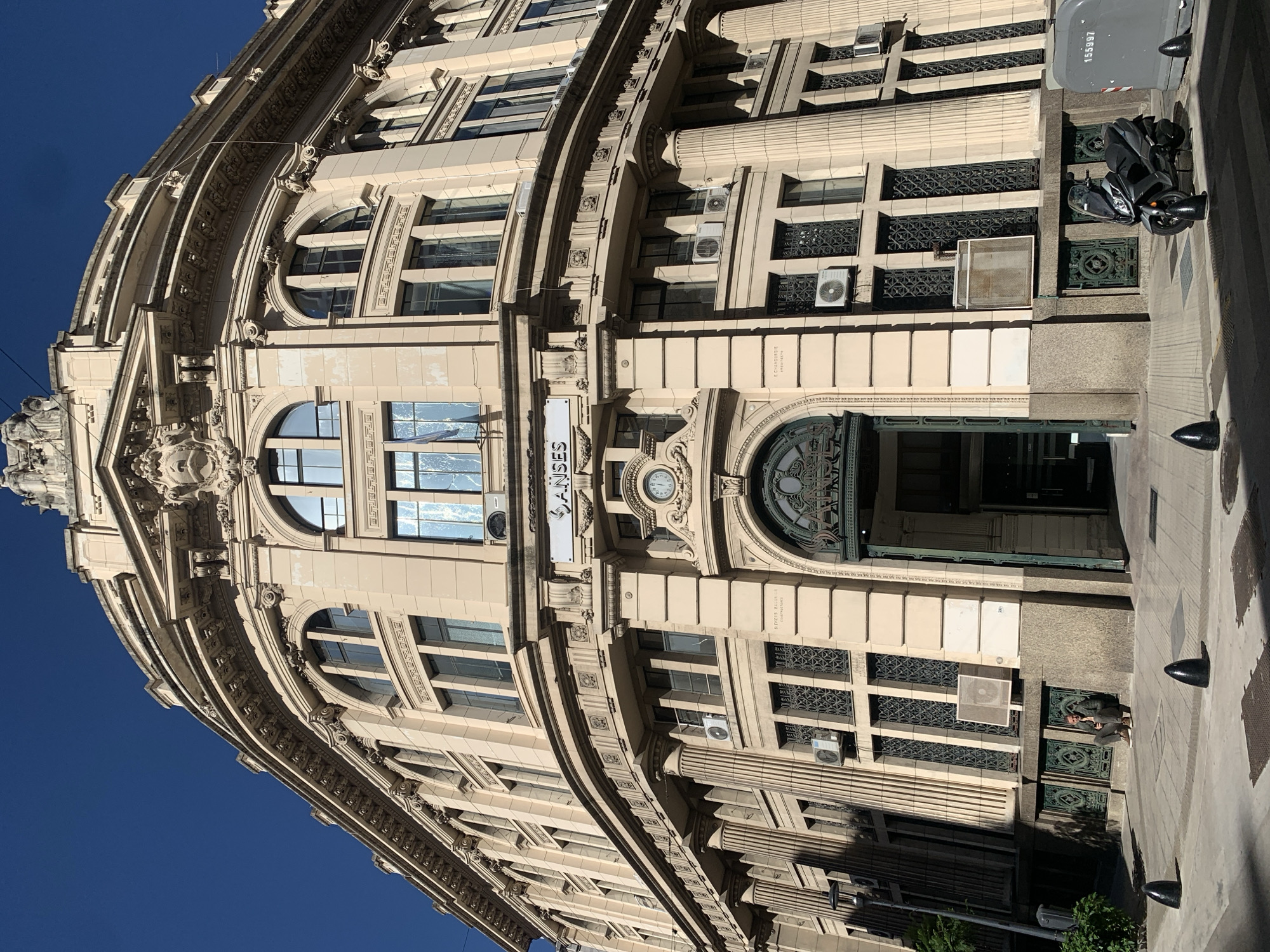 An ornate, cream-colored neoclassical or Beaux-Arts style building with a prominently curved facade dominates the scene under a clear blue sky. The multi-story structure features intricate architectural details, including engaged columns, decorative moldings, sculptures, and elaborate cornices. The lower floors display large arched windows and entrances, while upper floors have rectangular windows, many fitted with decorative dark iron grilles. Several modern white air conditioning units are mounted on the exterior walls.
Above the main arched entrance, a small circular clock is embedded in the facade, and a white rectangular sign reads "ANSES" in dark lettering. Another smaller sign on the right side of the main entrance facade reads "E. CURRODIGUE / ARQUITECTOS". The main entrance itself is grand, featuring an ornate dark metal gate or door.
On the paved sidewalk in front of the building, a black scooter or motorcycle is parked. Dark, rounded bollards are visible along the curb. In one cropped view, a person is seen sitting on the ground near the building. A gray utility box with the number "155997" is also present on the sidewalk. The scene is brightly lit by daylight, casting shadows that suggest a sunny day in an urban setting, consistent with Buenos Aires, Argentina.