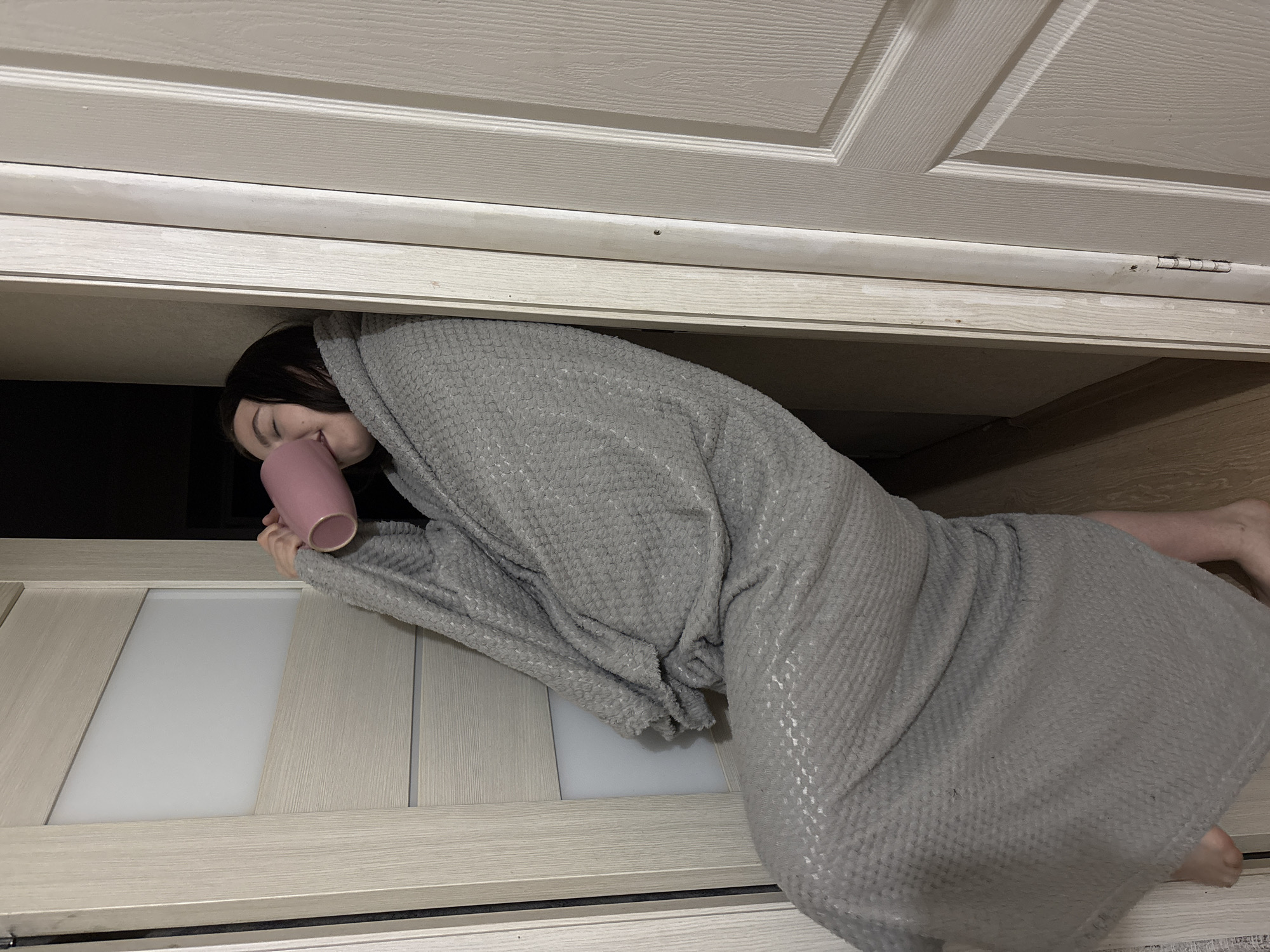 An indoor scene, possibly in Perm, Russia, depicts a person lying horizontally within a narrow, confined space, appearing to be between two doors or under a low shelf/cabinet. The image is oriented counter-clockwise, placing the "ceiling" to the left and the "floor" to the right.
The main subject is an individual, likely an adult female with dark hair, almost entirely covered by a thick, textured, light grey blanket or robe that has a waffle-like pattern. Her face is partially visible, showing her nose and mouth as she appears to be looking down or has her eyes closed. She holds a light pink or rose-colored mug in both hands near her face. Her bare feet are visible on the far right, resting on a light wooden surface, which appears to be a floor or a wide shelf.
Above the person (to the left in the rotated image) is a light-colored, paneled door or cabinet, possibly a closet door, with visible white hinges. Below the person (to the right) is another door, featuring a light wooden frame and a central frosted glass panel. The space the person occupies seems cozy and secluded. The lighting is generally dim, suggesting an indoor evening setting or a space with limited natural light. No text is visible in the image.