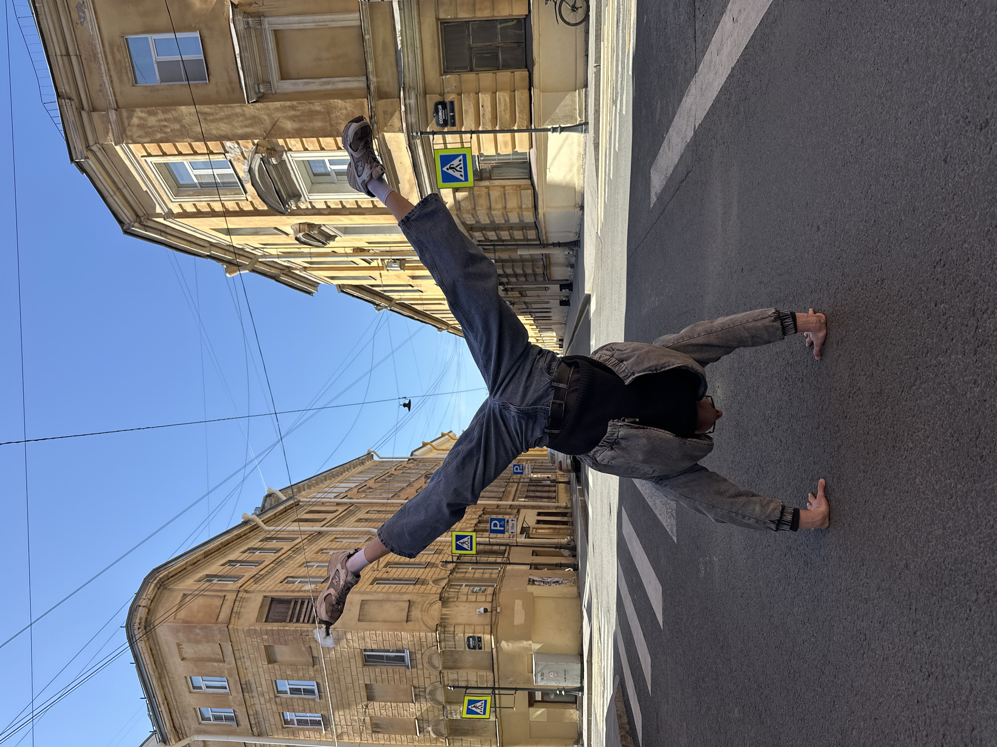 A person is captured in a dynamic pose, performing what appears to be a handstand or an inverted cartwheel in the middle of a paved street. They are dressed in dark-wash jeans, a black top, a grey or dark denim jacket, and light-colored athletic shoes with white socks, and wear glasses. Their body forms a wide 'V' shape, with legs extended towards the sky and hands firmly planted on the dark asphalt road.
The scene depicts an urban setting with historic architecture, likely in Saint Petersburg, Russia. Tall, ochre-colored buildings with multiple windows line both sides of the street, featuring classical architectural elements such as cornices and decorative window frames. The sky above is clear and bright blue, indicating sunny weather. Numerous utility wires crisscross the sky.
On the street, white pedestrian crosswalk markings are visible. Several street signs are present, including blue and white pedestrian crossing signs (a person walking) and blue parking signs. On a green street sign attached to a pole, the partial Russian word "САДОВАЯ" (Sadovaya), indicating a street name, is legible along with the number "1" below it. The low camera angle emphasizes the height of the buildings and the person's inverted position, creating a dramatic and energetic effect.