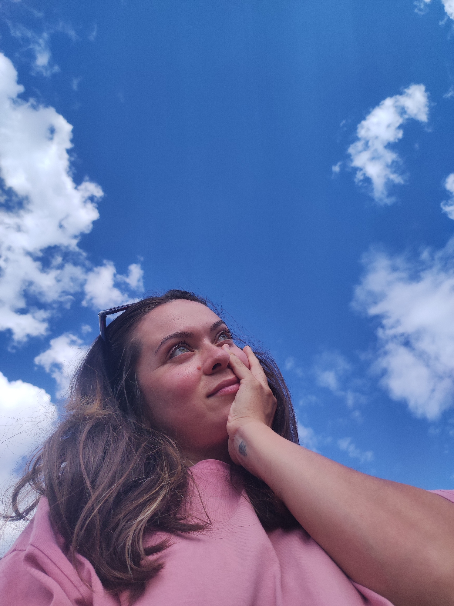 This is a low-angle, medium shot of a young woman looking up at a bright blue sky with scattered white clouds. She is wearing a pink crew-neck sweatshirt and has her right hand resting on her cheek. Her brown hair is styled loosely around her shoulders, and she has sunglasses perched on top of her head. A small tattoo is visible on her forearm. The lighting suggests it is daytime, likely mid-morning or afternoon, with clear weather. The woman's gaze is directed upwards, and her expression appears thoughtful or contemplative. The background is dominated by the sky, with no distinct location cues visible other than the implied outdoor setting. There is no visible text in the image.