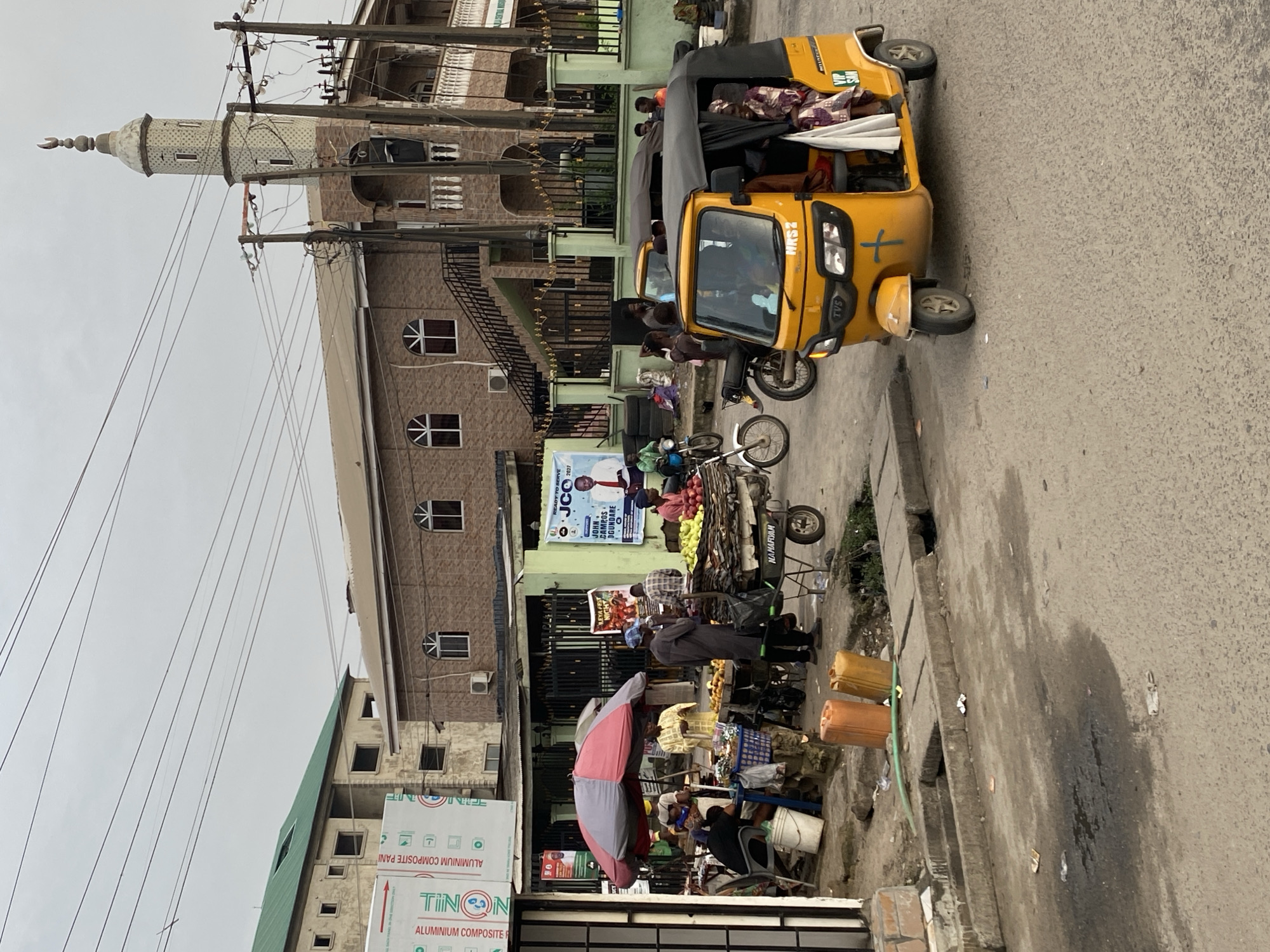 The image depicts a street scene in Ado, Nigeria, dominated by commercial activity and a yellow three-wheeled vehicle (a "keke"). A mosque with a distinct minaret is visible in the background. The street is lined with vendors and their stalls, selling fruits and other goods, with people interacting and shopping. In the foreground, various containers and a large umbrella are visible, further indicating market activity. A political campaign poster is also present, adding to the urban backdrop. Buildings of varied architectural styles are situated along the street, with electrical wires crisscrossing above.