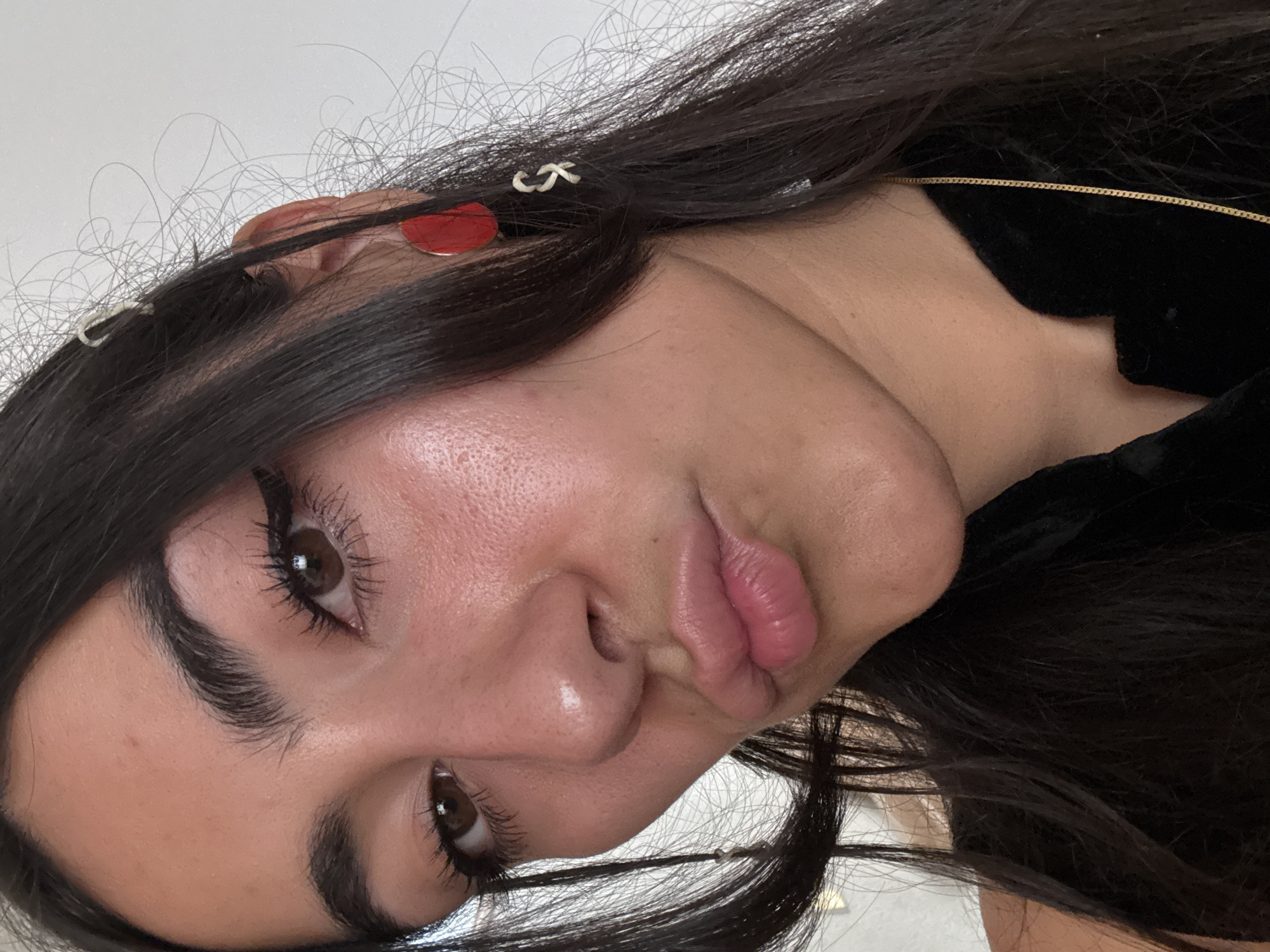 The primary subject of the image is a person, likely a young woman, captured in a close-up, diagonally oriented selfie. Her dark, long hair frames her face, with a red circular earring visible on her right ear and a small white hair clip subtly placed within her hair near the temple. A thin gold chain necklace is seen resting against the neckline of a black velvet garment.
The person is actively posing for the camera, looking directly into the lens with dark brown eyes. Her eyelashes appear enhanced with mascara, and her eyebrows are well-defined. Her lips are full and intentionally puckered into a "duck face" expression, conveying a playful or slightly pouty emotion. Her skin shows natural texture and a subtle glow. The background is a plain, light-colored surface, suggesting an indoor setting. While the image itself offers no specific location cues, the provided context indicates the location is Puebla, Mexico. There is no discernible text within the image.