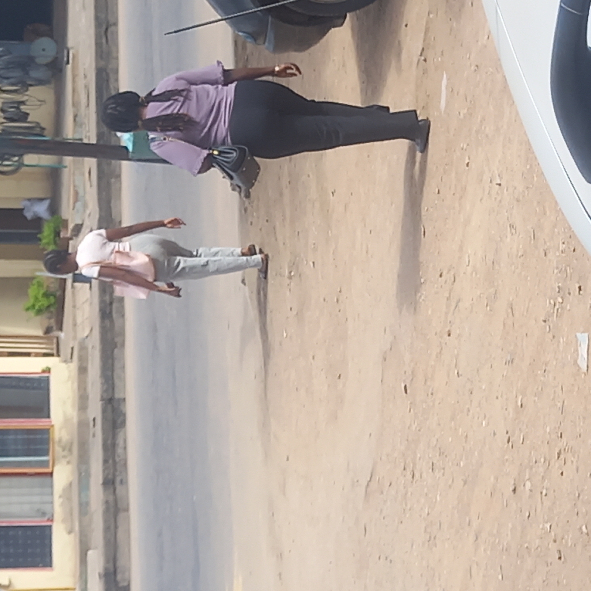 Two women are walking on a street in Dadi Riba, Nigeria. One woman is wearing a purple shirt and black pants with a black purse, while the other is wearing a pink shirt and gray pants. A car is parked on the right side of the image, and buildings are visible on the left. The ground appears to be dirt or unpaved.