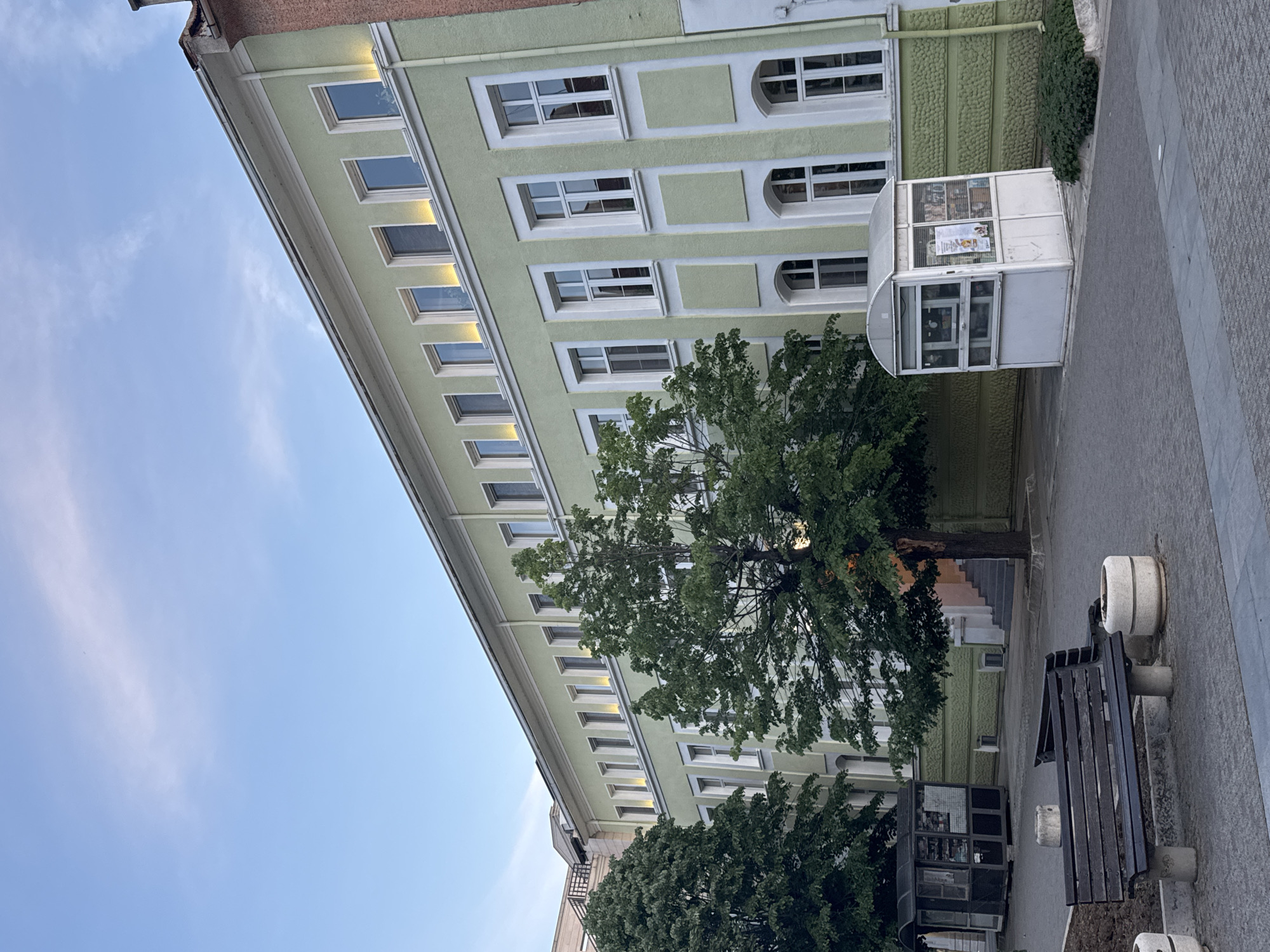 The image shows a multi-story, light green building with white-framed windows. A tree with green foliage partially obscures the building. A small white kiosk stands in front of the building. A bench and cylindrical concrete planters are visible on the right side of the image. The sky is visible in the upper left corner. The location is likely in a city, but the specific city is unknown. The scene appears to be outdoors, likely in a courtyard or public space.