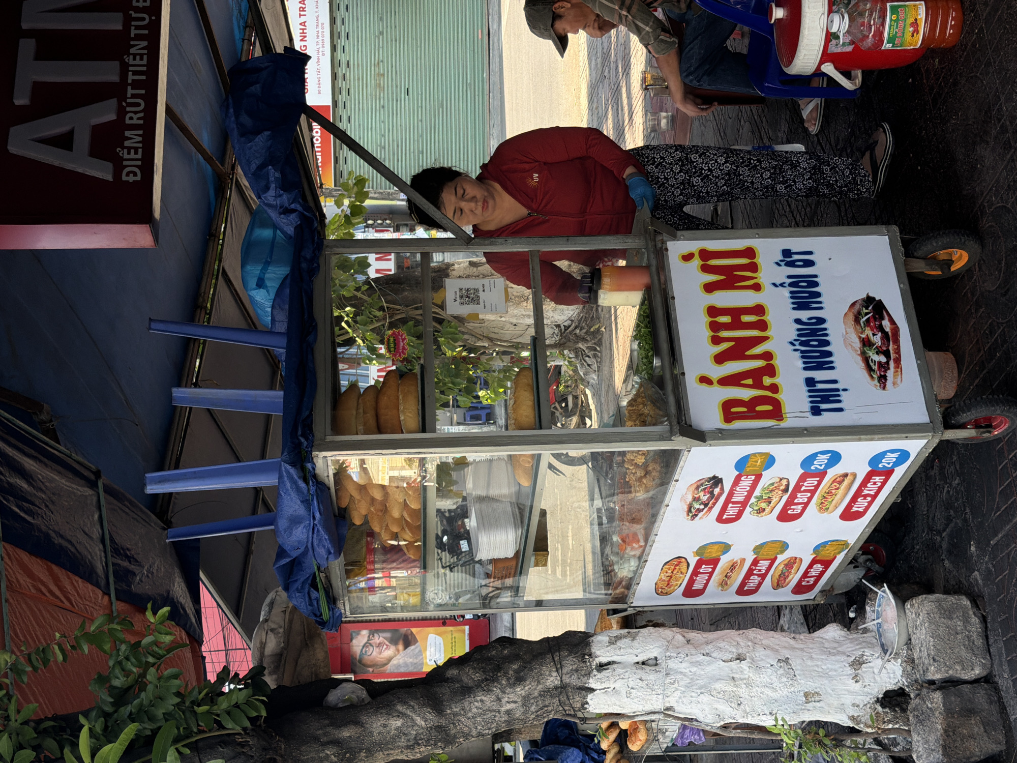 A woman in a red zip-up jacket and patterned pants prepares food at a mobile bánh mì stand in Bac Nha Trang Ward, Vietnam. The stand has a glass display case filled with bread and ingredients. A sign on the stand advertises various bánh mì fillings. A man in a cap sits on a blue chair to the right of the stand, and a large red cooler is beside him. The scene takes place outdoors on a paved surface, with a tree and a building in the background.