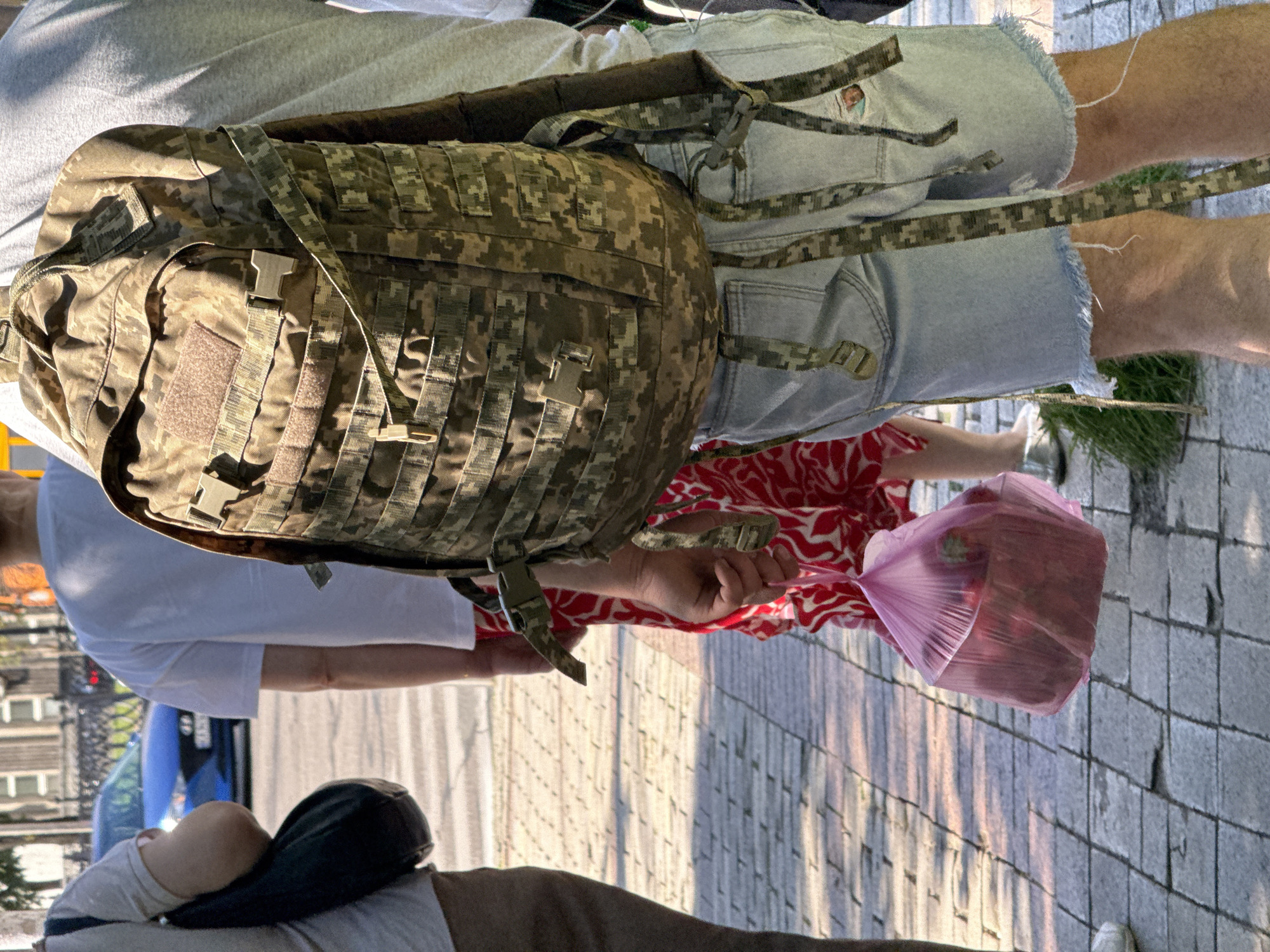The image shows a close-up of a person from the waist down wearing light blue denim shorts with a frayed hem and a large, camouflage-patterned backpack. The backpack is a tactical style with multiple straps and buckles. The person is standing on a sidewalk with a patterned stone surface. In the background, other people are visible, including one wearing a red and white patterned dress and holding a pink plastic bag filled with red items, possibly fruit. Another person in the background is wearing a white t-shirt. A blue car is also visible on a street behind the people. The scene appears to be outdoors in an urban environment, likely a city street or square. The lighting suggests it is daytime. Based on the specific camouflage pattern on the backpack and the presence of what appears to be the Ukrainian flag on a patch on the backpack, this scene is likely in Kyiv, Ukraine.