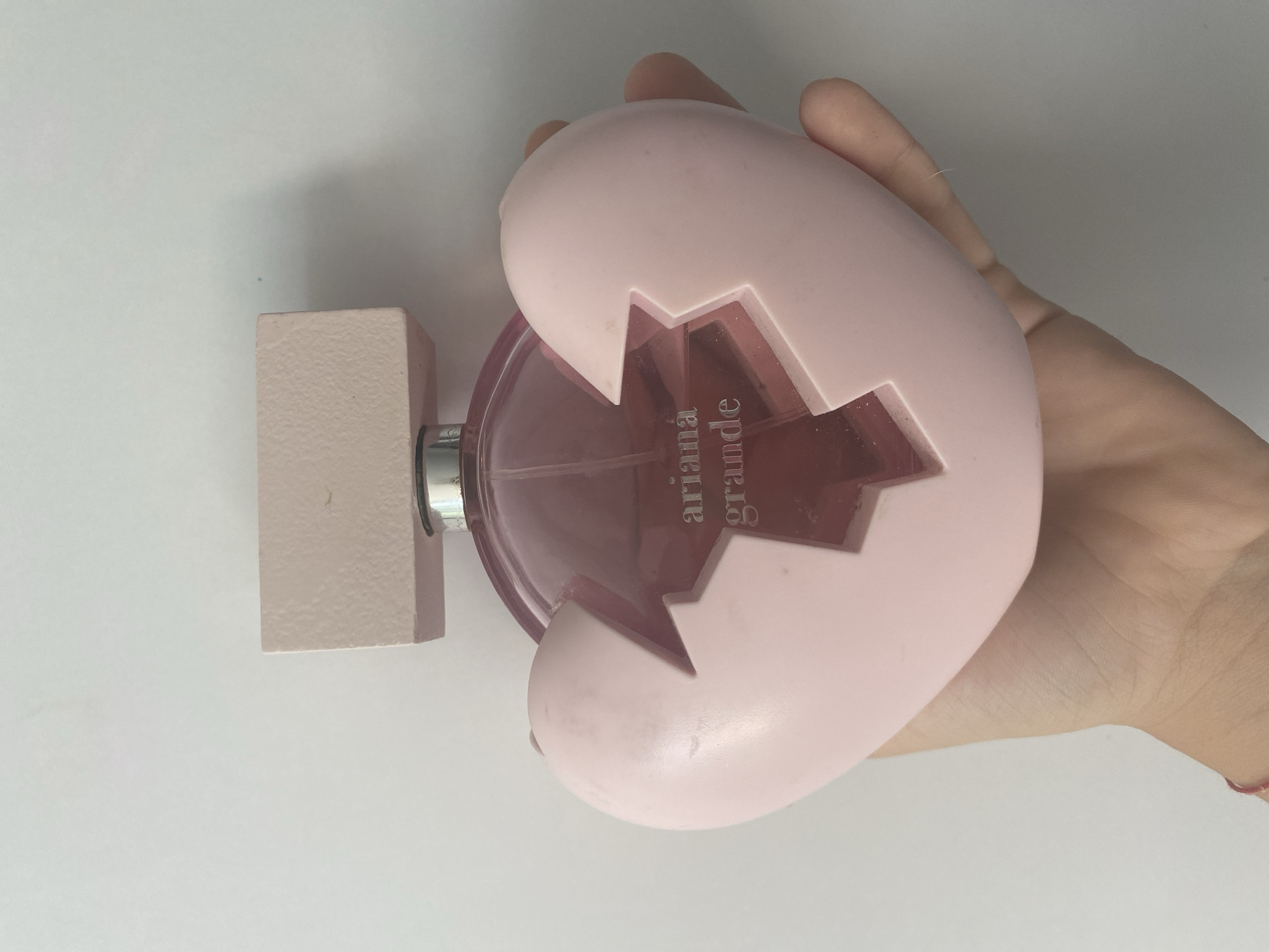A light pink perfume bottle, shaped like a broken heart, is held in a person's left hand against a white background. The bottle has a rectangular cap. The brand "Ariana Grande" is embossed in silver on the glass portion of the bottle. The scene is indoors.