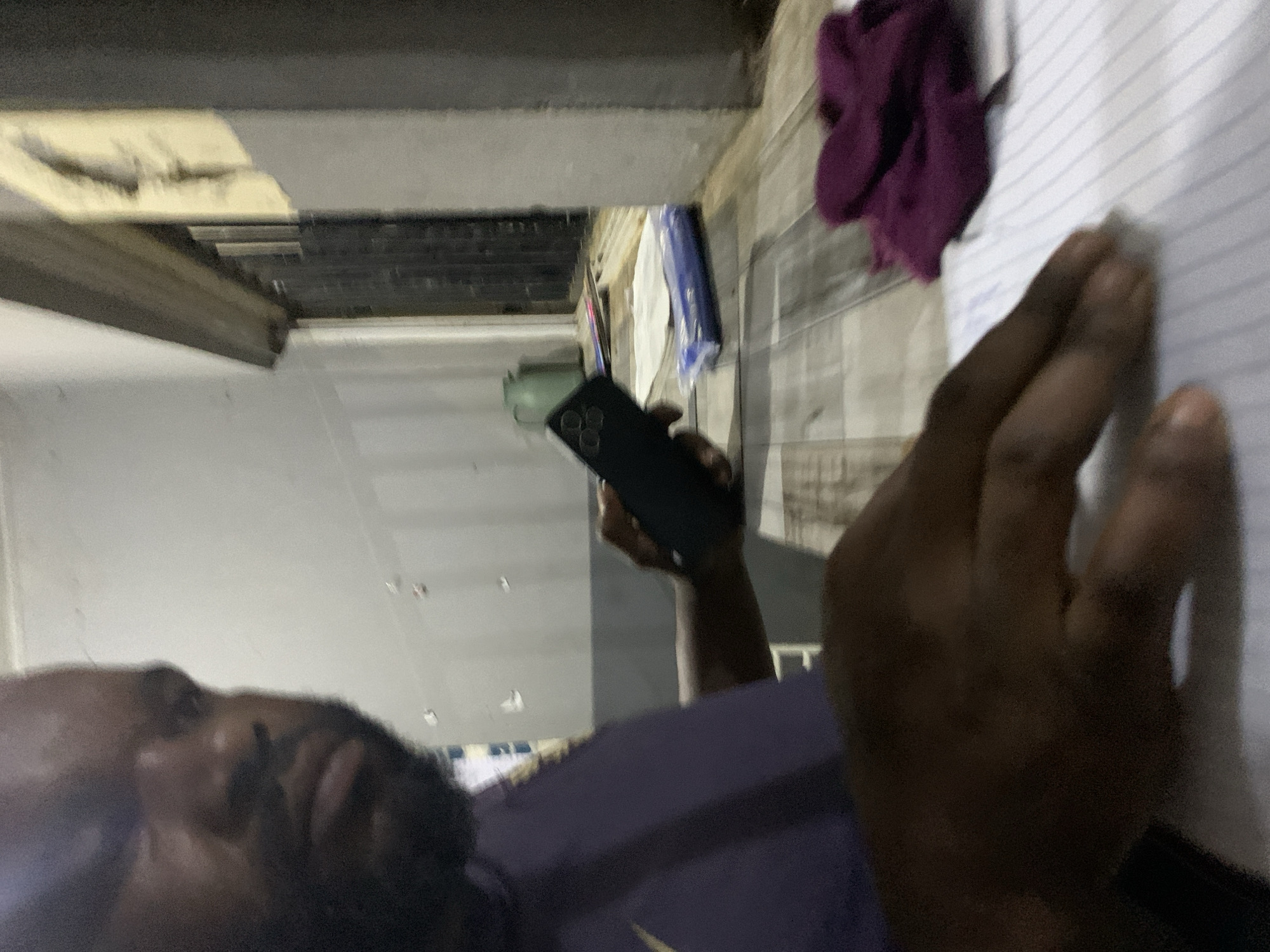 The scene, located in New Stadium, Nigeria, depicts a dark-skinned individual with a beard and close-cropped dark hair or a bald head, positioned indoors. The person, wearing a purple short-sleeved shirt with a light-colored logo on the left shoulder, is partially visible from the chest up on the left side of the frame, looking downward and to their right. Their left hand holds a dark smartphone featuring a prominent triple-lens camera array on its back. Their right hand rests on a white sheet of lined paper, which is placed on a light-colored surface with a distinct wooden plank pattern. To the right of the paper, a crumpled purple textile object is present. In the mid-ground, a dark, slatted window or service opening with a light frame is visible. Behind this opening, a small green pitcher and additional stacked papers are situated on a ledge. The background wall is light-colored and exhibits several small, irregularly shaped dark marks. The environment is illuminated by artificial light.