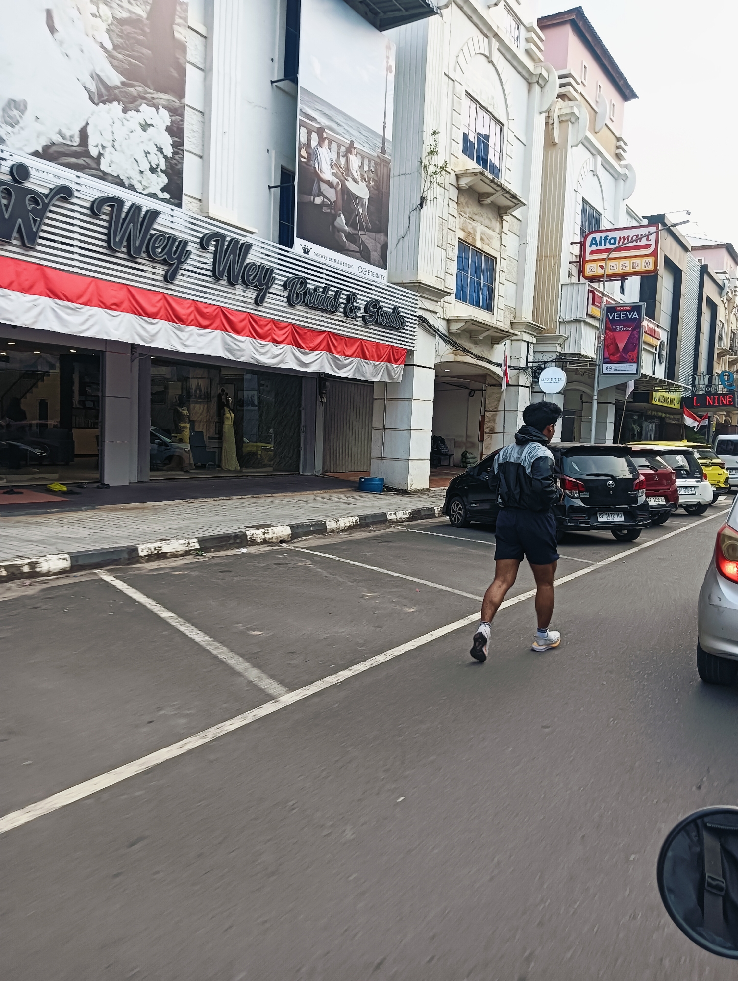 A young man with dark, curly hair and wearing a grey and black jacket, dark blue shorts, and athletic shoes is walking across a street in a city. He is facing away from the camera. The street has visible white parking lines. To his left is a sidewalk with a building facade that features large banners with images of wedding dresses and couples. The building also has signage that reads "Wey Wey Bridal & Studio." Further down the street, other buildings are visible, including an "Alfamart" convenience store and a store with a "VEEVA" advertisement. Several cars are parked along the street, including a black sedan, a red sedan, and a yellow sedan. The sky is overcast. The perspective of the image suggests it was taken from a moving vehicle, possibly a motorcycle, as a circular mirror is partially visible in the lower right corner.