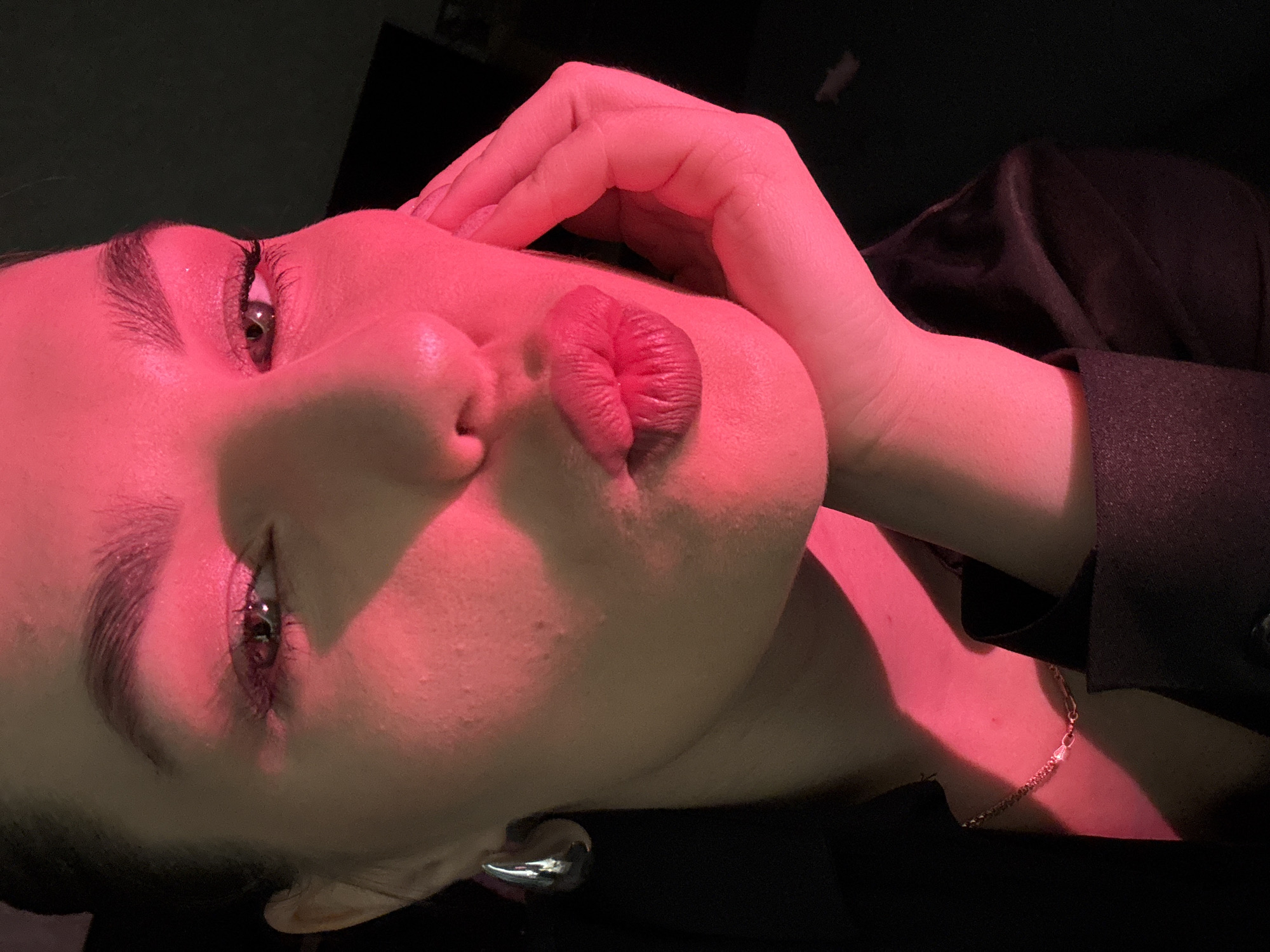 A close-up, medium shot captures a woman's face illuminated by pink light against a dark background. She is angled towards the viewer, her lips pursed and slightly open, with her right hand gently touching her cheek. Her eye makeup features pink eyeshadow and dark mascara. She wears a dark jacket and a delicate necklace. A large, silver, tear-drop shaped earring is visible on her left ear. The scene appears to be indoors, possibly a studio or a dimly lit room. There is no discernible connection to the city of Chaikovsky, Russia.