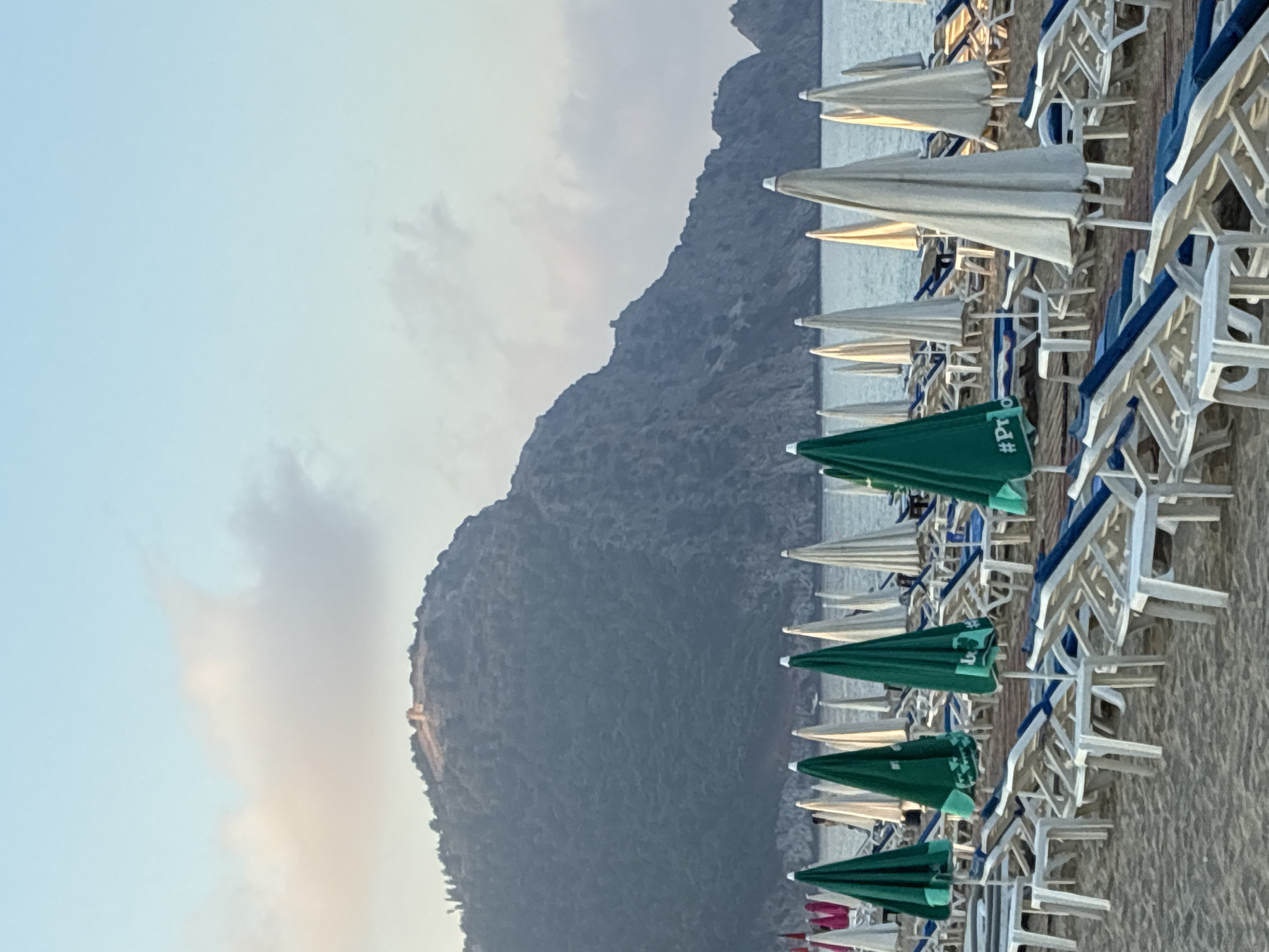 The image depicts a beach scene in Alanya, Turkey, with numerous empty lounge chairs and closed umbrellas arranged on the sand. In the background, a large, rugged mountain with a structure on its summit dominates the landscape, overlooking the calm sea. The sky is a pale blue with soft, wispy clouds. The overall impression is one of a serene, early morning or late afternoon setting on a popular tourist beach.