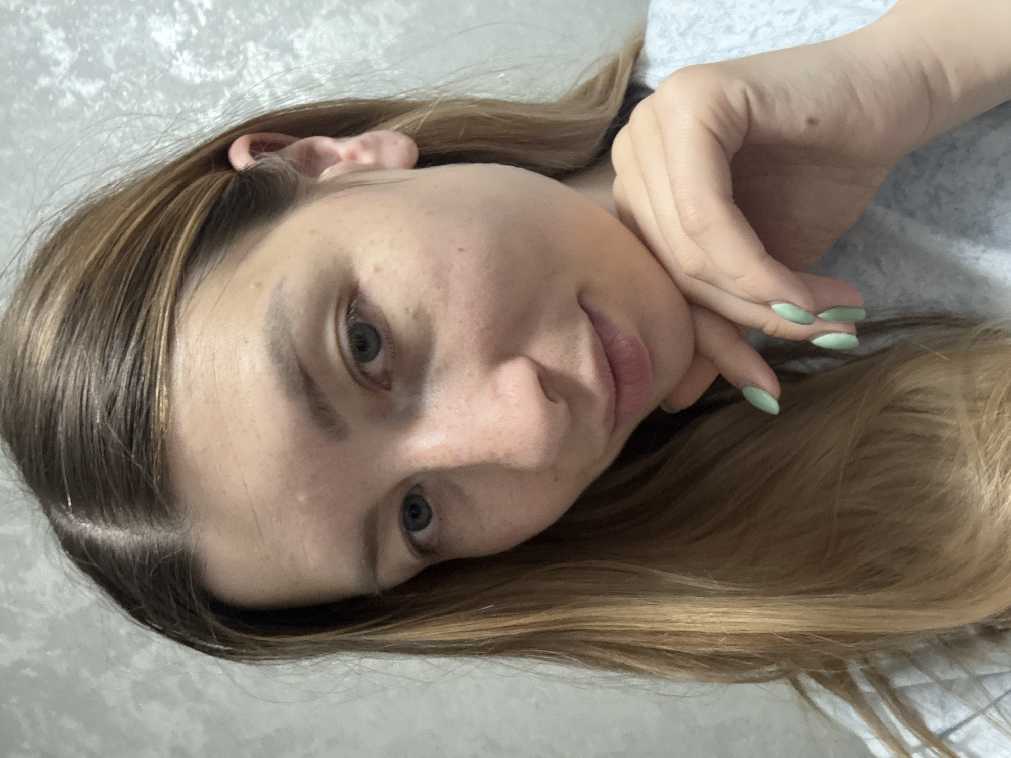 A young woman with long, light brown hair is lying down, her head resting on a gray surface that appears to be a bed or couch. Her face is turned towards the camera, and her eyes are open. She has fair skin with some freckles and blemishes. Her right hand is raised near her face, with her fingers curled slightly. She has a light green polish on her nails. The background is a textured gray surface. The image does not show the location in Moscow, Russia.