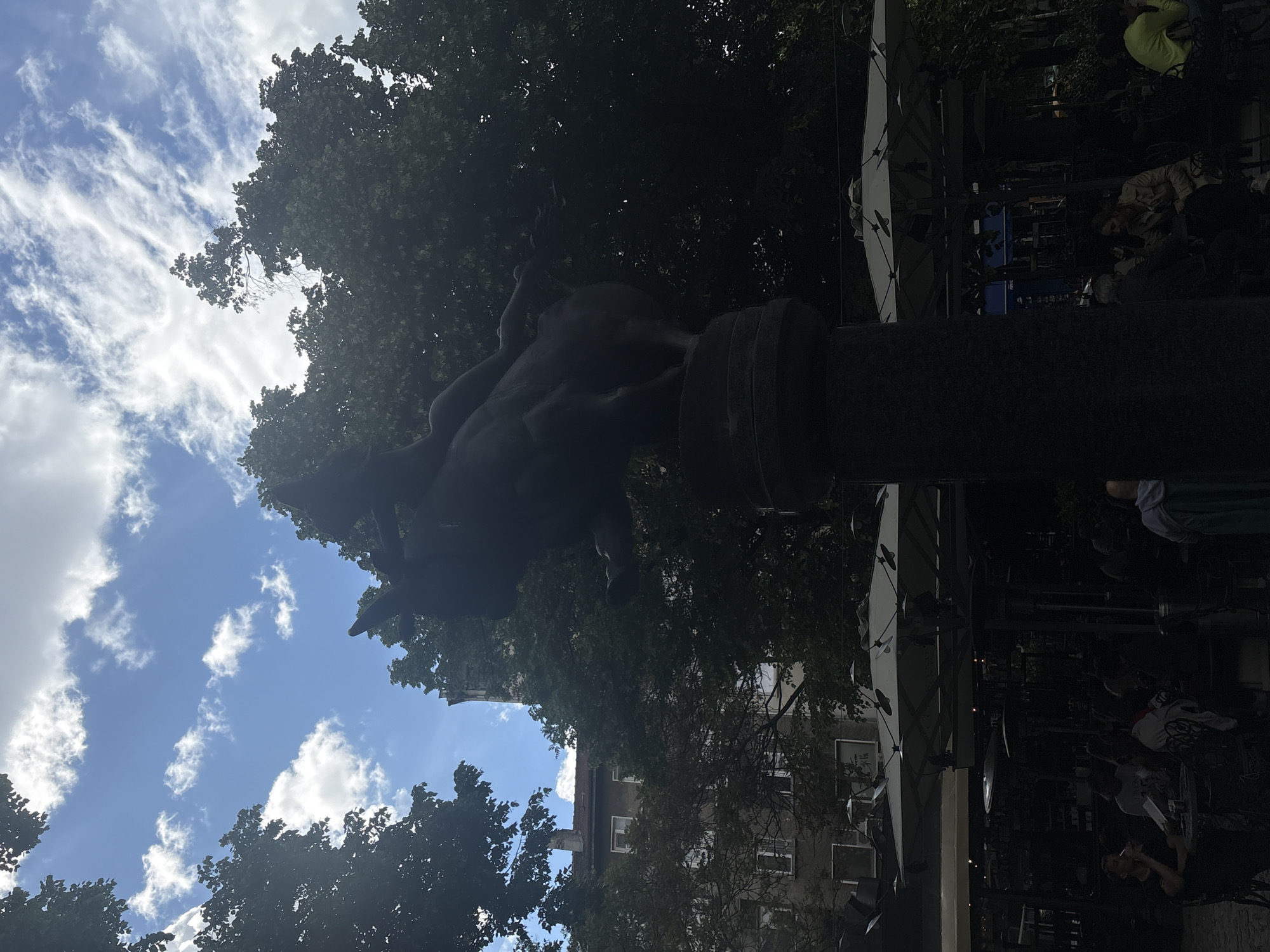 The image shows a statue atop a pillar in what appears to be a city square. The statue is dark and indistinct, but it seems to depict figures in motion, possibly a human riding an animal. Surrounding the statue are trees and buildings, with a bright sky visible in the background featuring scattered clouds. The base of the pillar is also dark, obscuring any details. To the right of the statue, there is an outdoor seating area with tables and people. The overall impression is that of an urban landscape with historical and modern elements.
