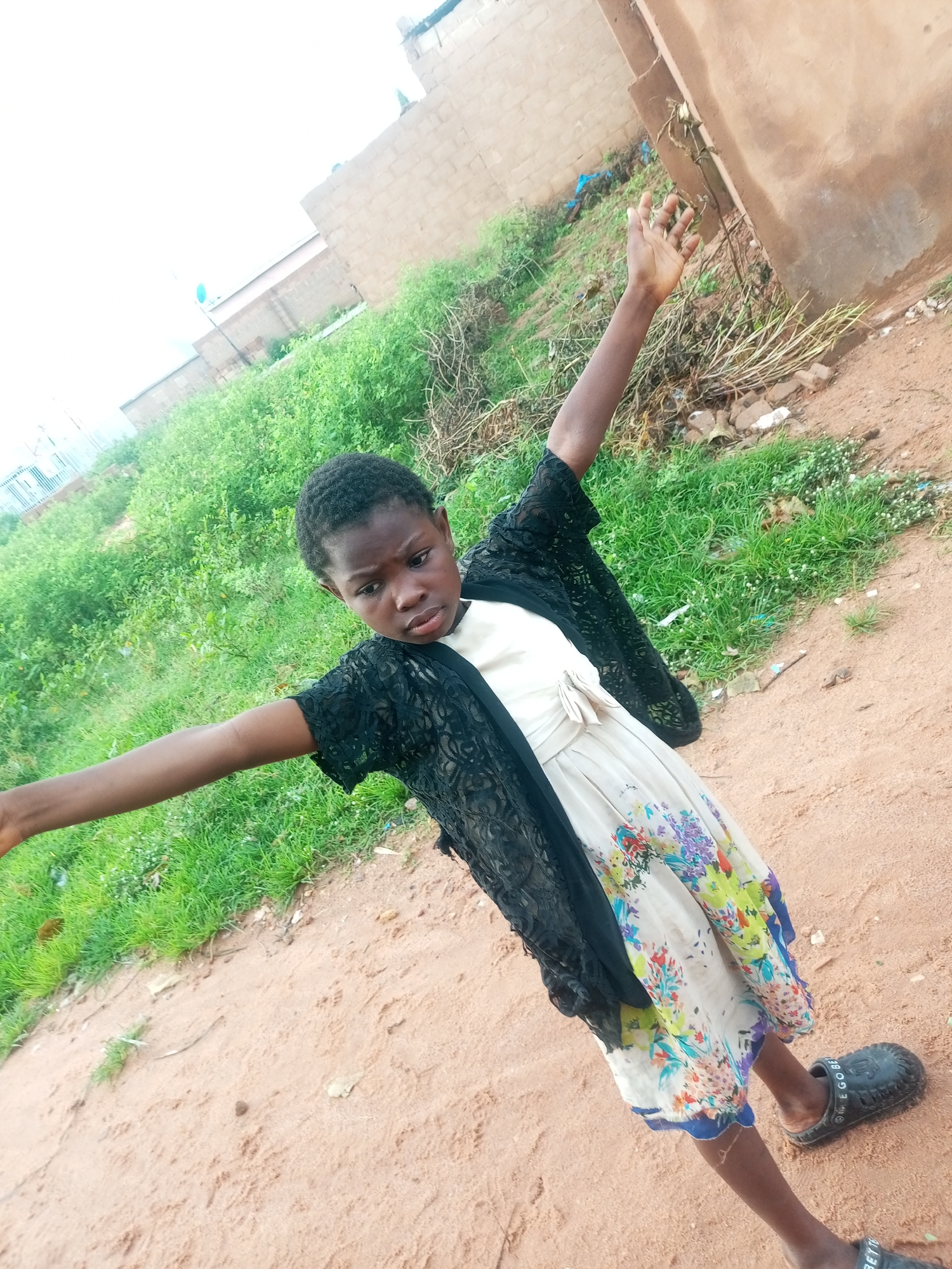 A young Nigerian girl stands outdoors on a dirt path in Tempure, Nigeria. She is wearing a cream-colored dress with a floral pattern, a black lace cardigan, and dark sandals. Her arms are outstretched, and she is looking to her left. The background consists of green foliage, a beige brick building, and a brown wall.