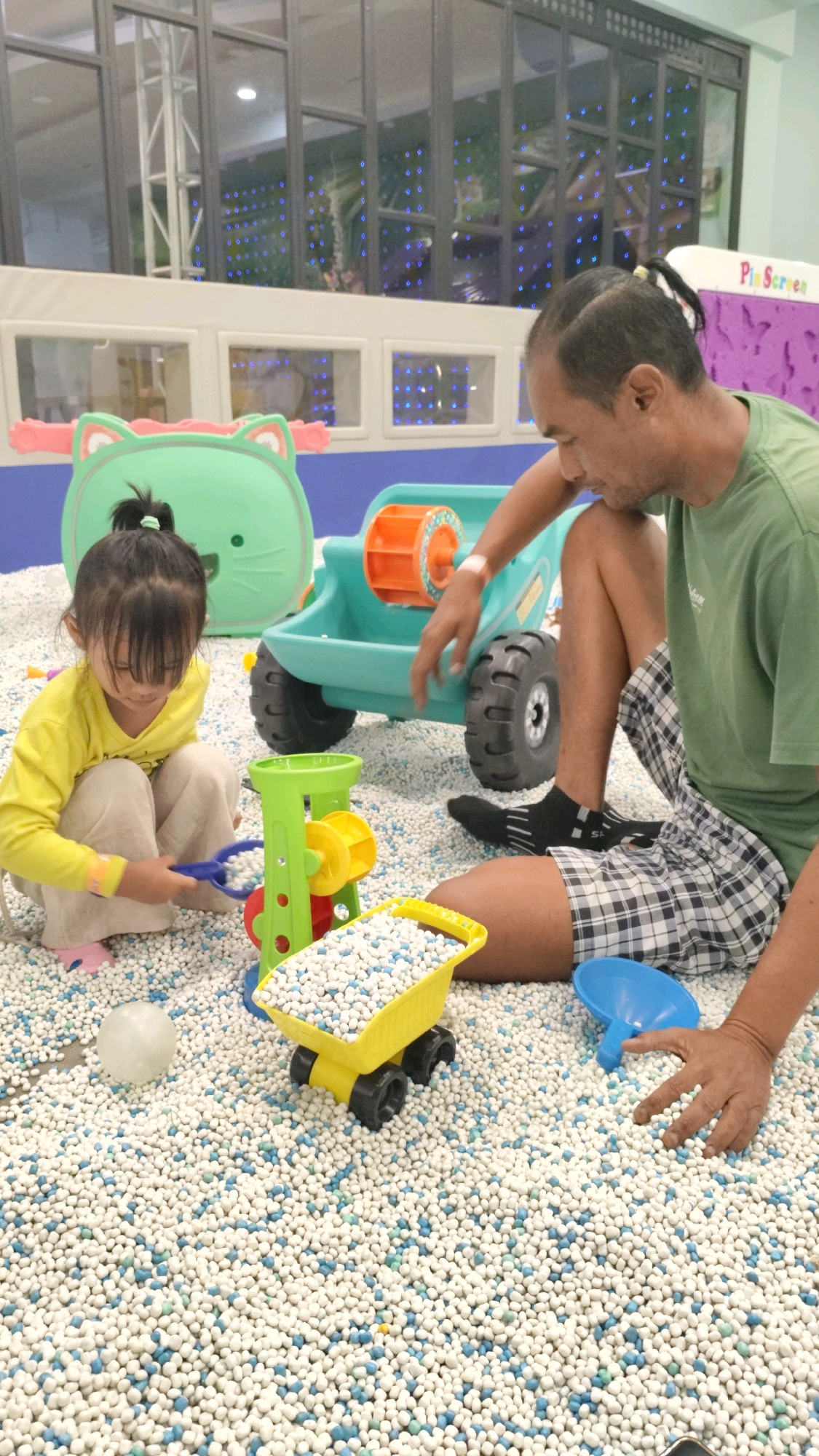 In an indoor play area filled with small white and blue balls, a young girl wearing a yellow long-sleeved shirt and beige pants is kneeling and playing with a blue plastic toy. Beside her is a green toy crane with yellow and red components. To her right, a man in a green t-shirt and plaid shorts is sitting and interacting with a teal toy truck. The background shows a large green cat-shaped toy. The overall environment appears to be a child-friendly indoor entertainment facility, possibly located in Bandung City, Indonesia, given the context of the query.