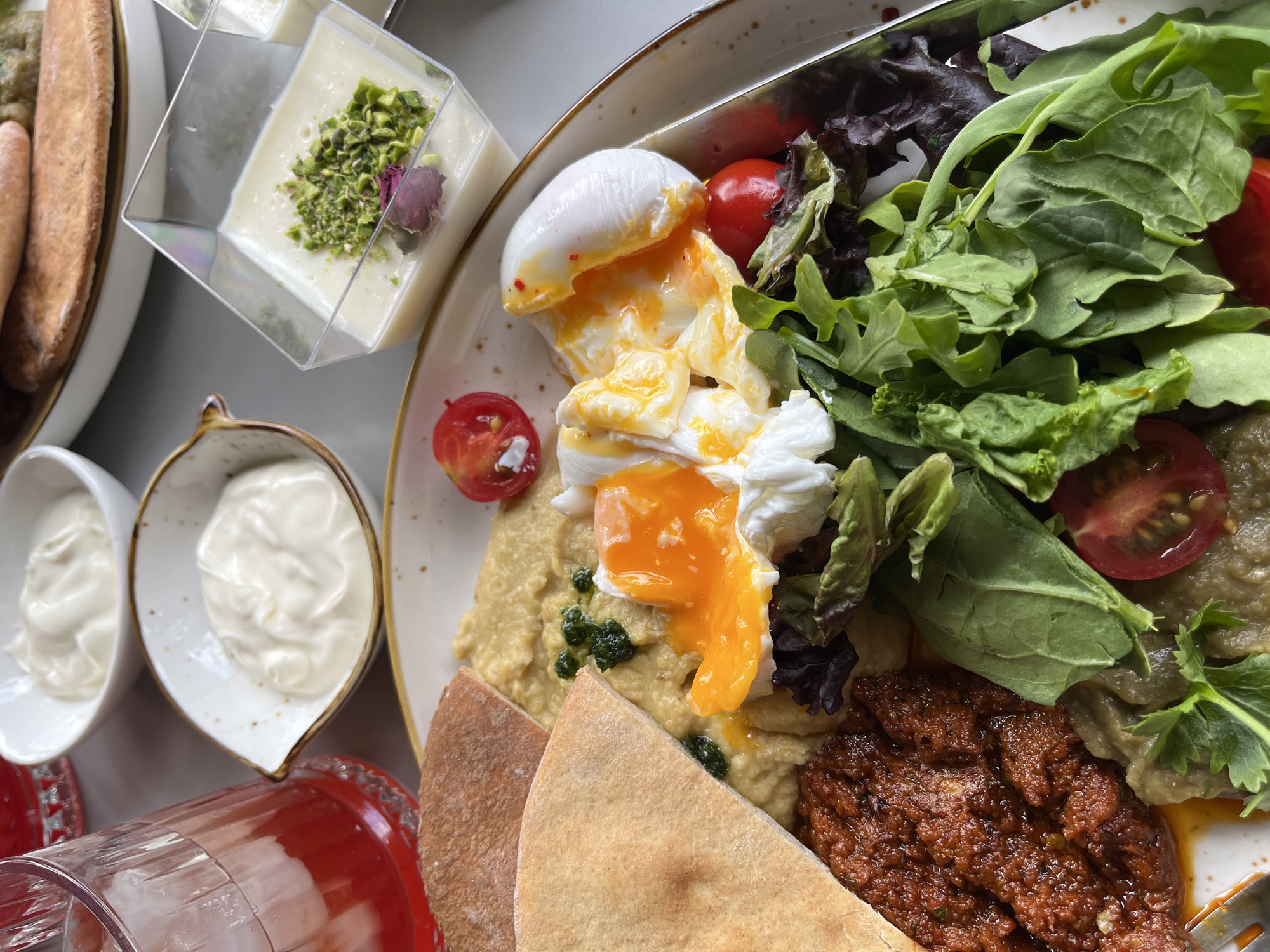 A close-up shot displays a breakfast spread, likely served at a cafe or restaurant, with the city of Almaty, Kazakhstan, in the background. In the foreground, a plate holds a dish featuring a perfectly poached egg with a runny yolk, nestled amongst a bed of mixed greens and halved cherry tomatoes. Alongside, there's a portion of hummus, a dollop of a dark red spread, and pieces of pita bread. In the background, several small bowls are visible: one containing a white creamy substance, another holding a portion of what appears to be yogurt with pistachios and a rose petal garnish, and a third with more of the creamy white substance. A glass of vibrant red liquid, possibly a beverage with ice, is also present. The setting suggests a leisurely breakfast meal, with the subtle hints of Almaty's cityscape implying an urban dining experience.