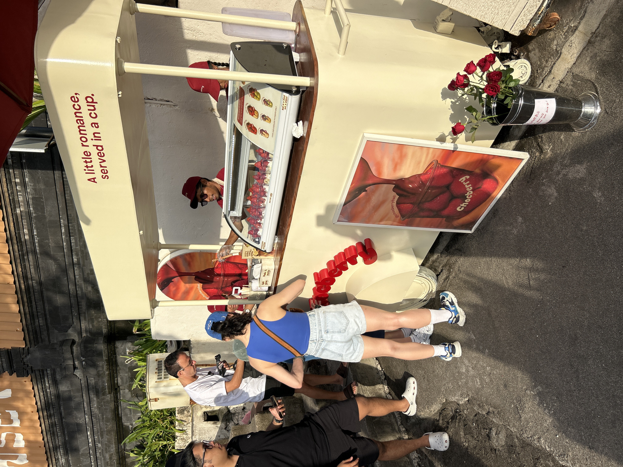 This is a medium-shot photograph taken outdoors in the city of North Kuta, Indonesia, depicting a scene in front of a dessert stand. The stand itself is cream-colored with a red awning and features a glass display case filled with strawberry desserts. Above the display, the text "A little romance, served in a cup" is visible in red lettering. Two individuals, wearing red caps and what appear to be uniform shirts, are working behind the counter.
In front of the stand, four people are present. On the left, a man in a black polo shirt and black shorts holds a smartphone. Next to him, a man in a white t-shirt and black shorts is taking a picture with a camera. In the center, a woman wearing a blue tank top, light-wash denim shorts, white socks, and athletic shoes stands facing the dessert stand. Her left hand is reaching towards the display. To her right, another person's legs and feet are visible. A large framed poster displaying a chocolate-covered strawberry dessert is leaning against the stand. To the right of the poster, a silver vase holds a bouquet of red roses. The ground is paved with rough, dark asphalt. The lighting suggests it is daytime, possibly late afternoon, given the long shadows.