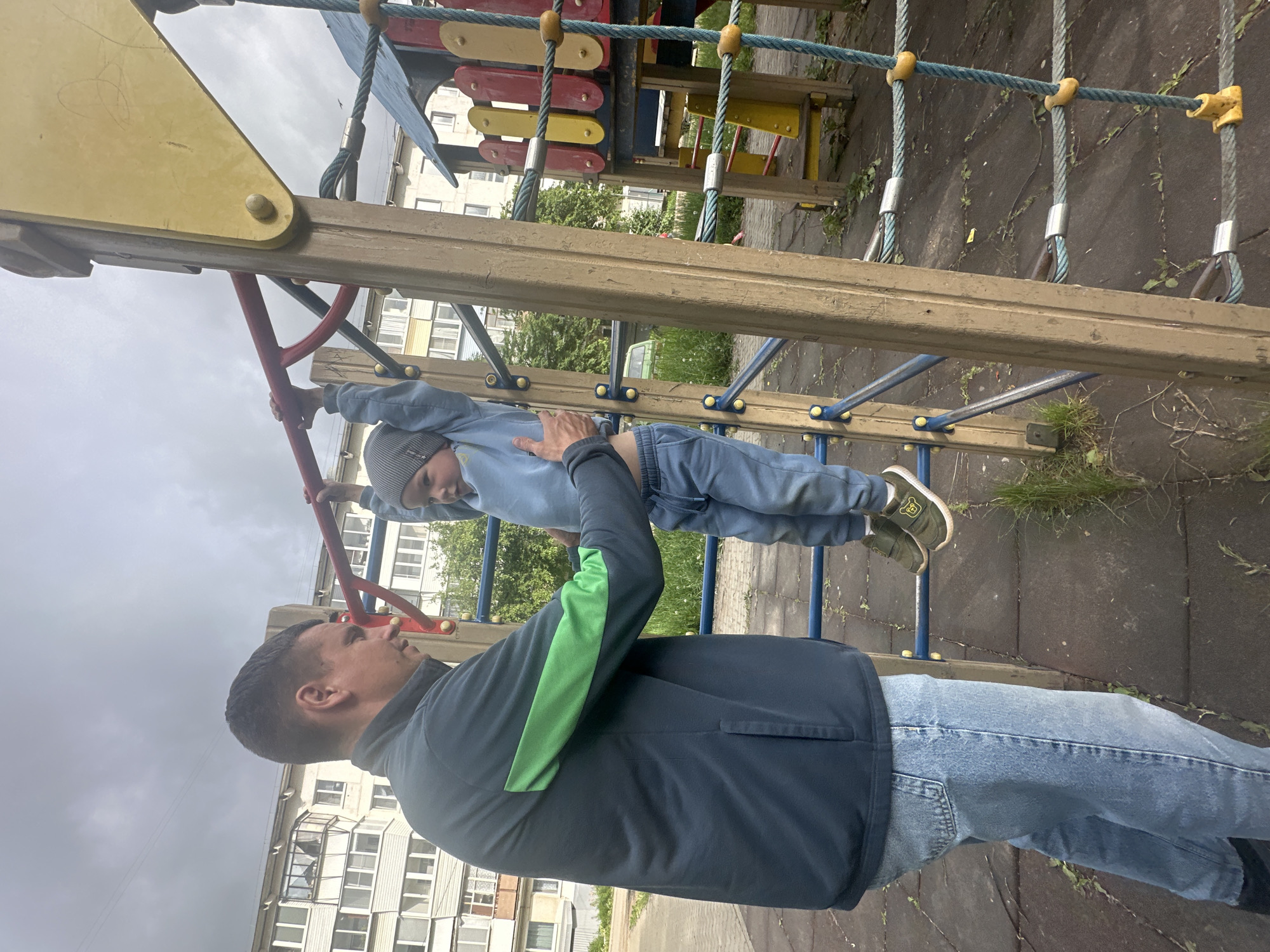 An outdoor scene in Chaikovsky, Russia, shows a man assisting a young child on playground equipment under an overcast sky. The child, dressed in a light blue sweatsuit, a striped beanie, and olive green sneakers with a yellow sole and a monkey emblem, is hanging from a red metal bar, seemingly supported by the man's hand on their lower back. The child looks focused or slightly straining.
The man, viewed from the side, wears a dark blue zip-up jacket with a bright green stripe on the back of his left sleeve, light blue jeans, and dark shoes. He is looking up at the child with an attentive expression.
The playground features various elements, including a yellow, triangular panel, red and yellow wooden planks, blue horizontal bars, and a rope climbing net with blue ropes and yellow plastic connectors. In the background, several multistory residential buildings with multiple windows are visible. The ground beneath the playground is a mix of paved areas, dirt, and sparse patches of grass and weeds. The sky is uniformly gray and cloudy, suggesting an overcast daytime. No visible text is present in the image.