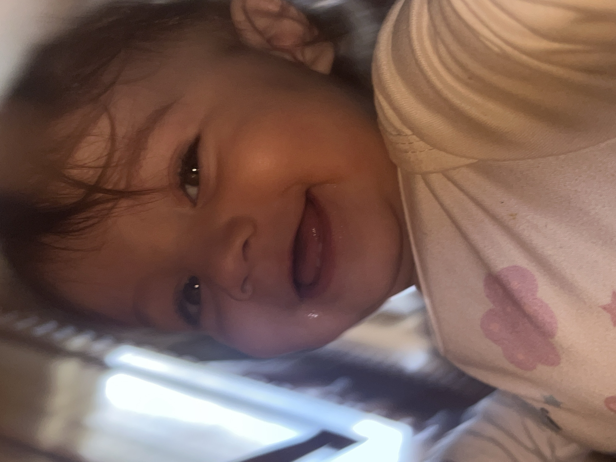 A baby lies sideways, resting on a surface with patterned edges. The baby wears a light-colored garment with a pink cloud design. The baby is smiling, with a small bubble of drool visible near their mouth. The location and city are not discernible from the image.