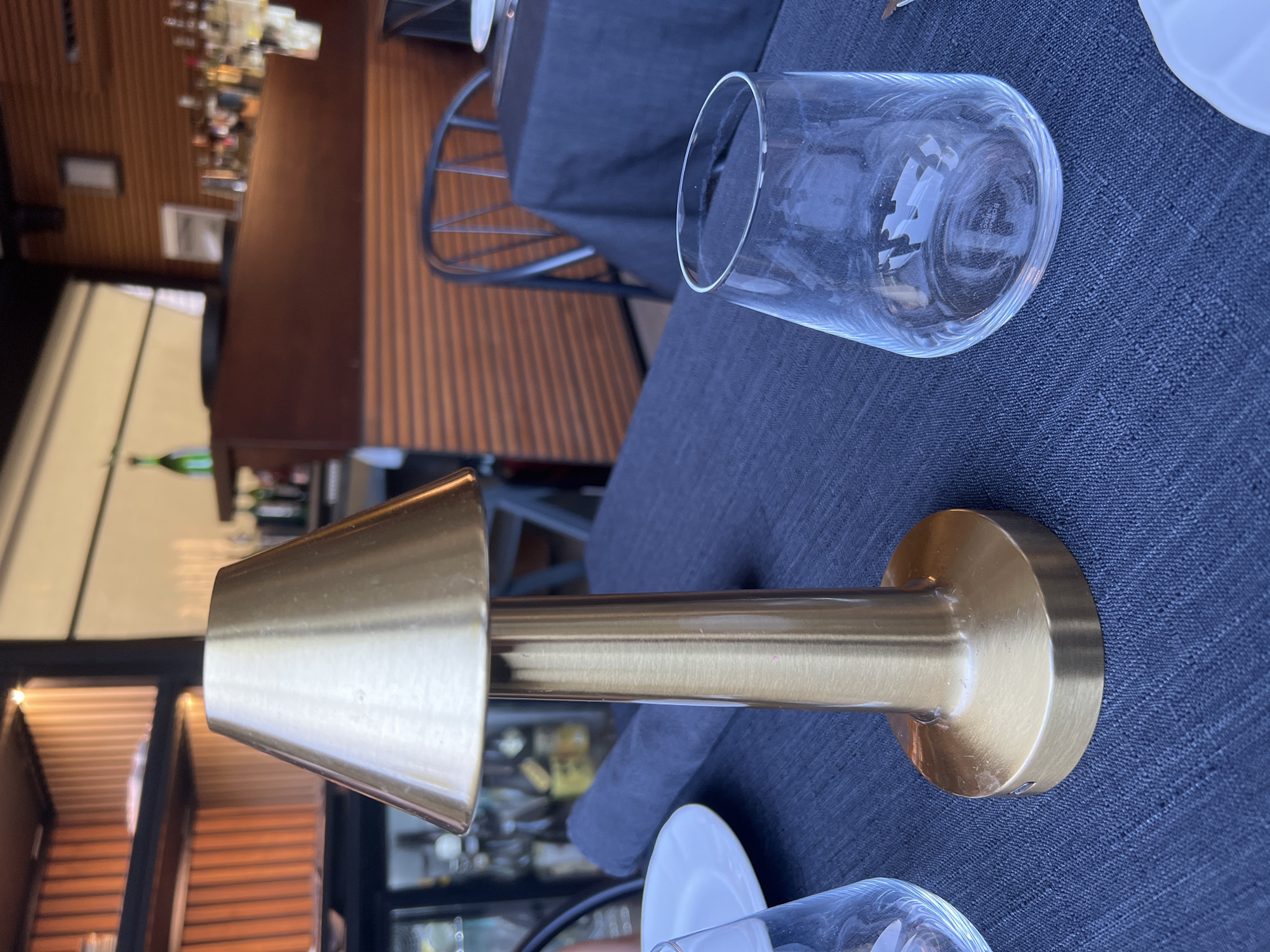 The image depicts a close-up view of a table setting at what appears to be a restaurant. The foreground is dominated by a brass table lamp with a conical shade, placed on a dark blue tablecloth. To the left of the lamp, a white plate is visible, partially obscured by a clear glass. To the right of the lamp, another clear glass, possibly a wine glass, is shown. The background reveals shelves lined with what seem to be bottles and decorative items, suggesting a bar area. A wooden-paneled wall and part of a chair are also visible, indicating an indoor or covered outdoor dining space. The scene is well-lit, with the lamp and overhead lighting contributing to the ambiance. There are no people directly visible in the frame. The setting appears neat and prepared for dining. There is no indication of the city of Rome, Italy.