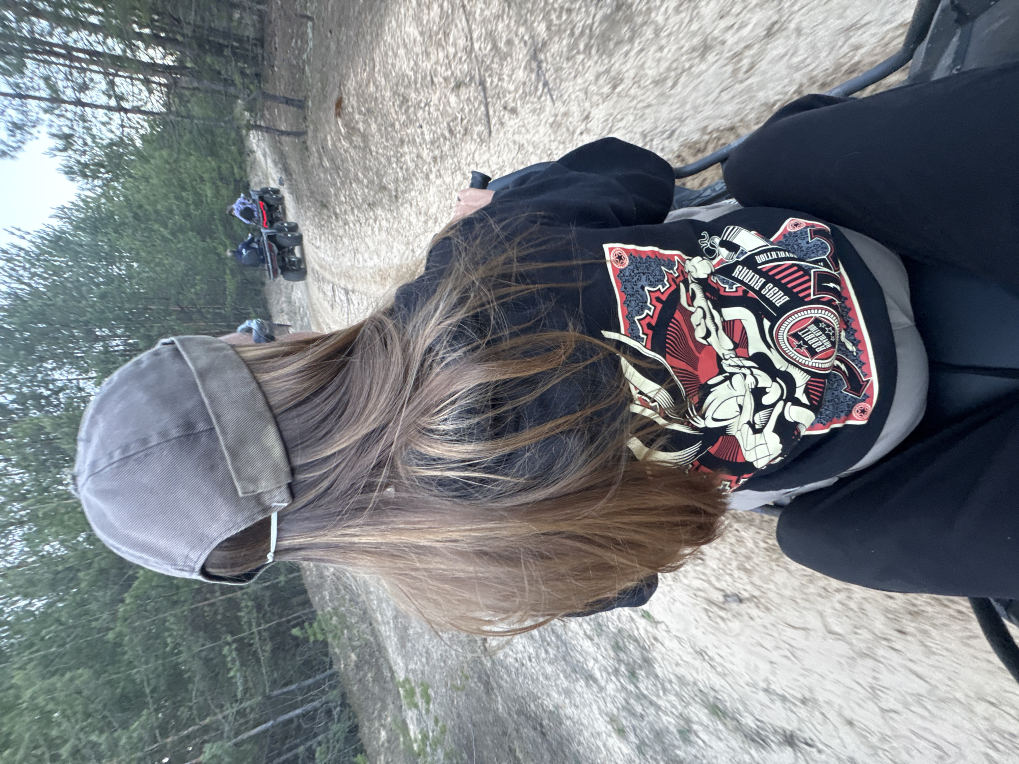 A person wearing a gray baseball cap, black pants, and a black hoodie with a distinctive graphic on the back is riding on a four-wheeled vehicle. The ground is light-colored, suggesting a sandy or dusty terrain. Trees are visible in the background, indicating a wooded area. Two other four-wheeled vehicles are visible further down the path. The location appears to be in a natural or semi-rural environment in Bol'shoye Kozino, Russia.