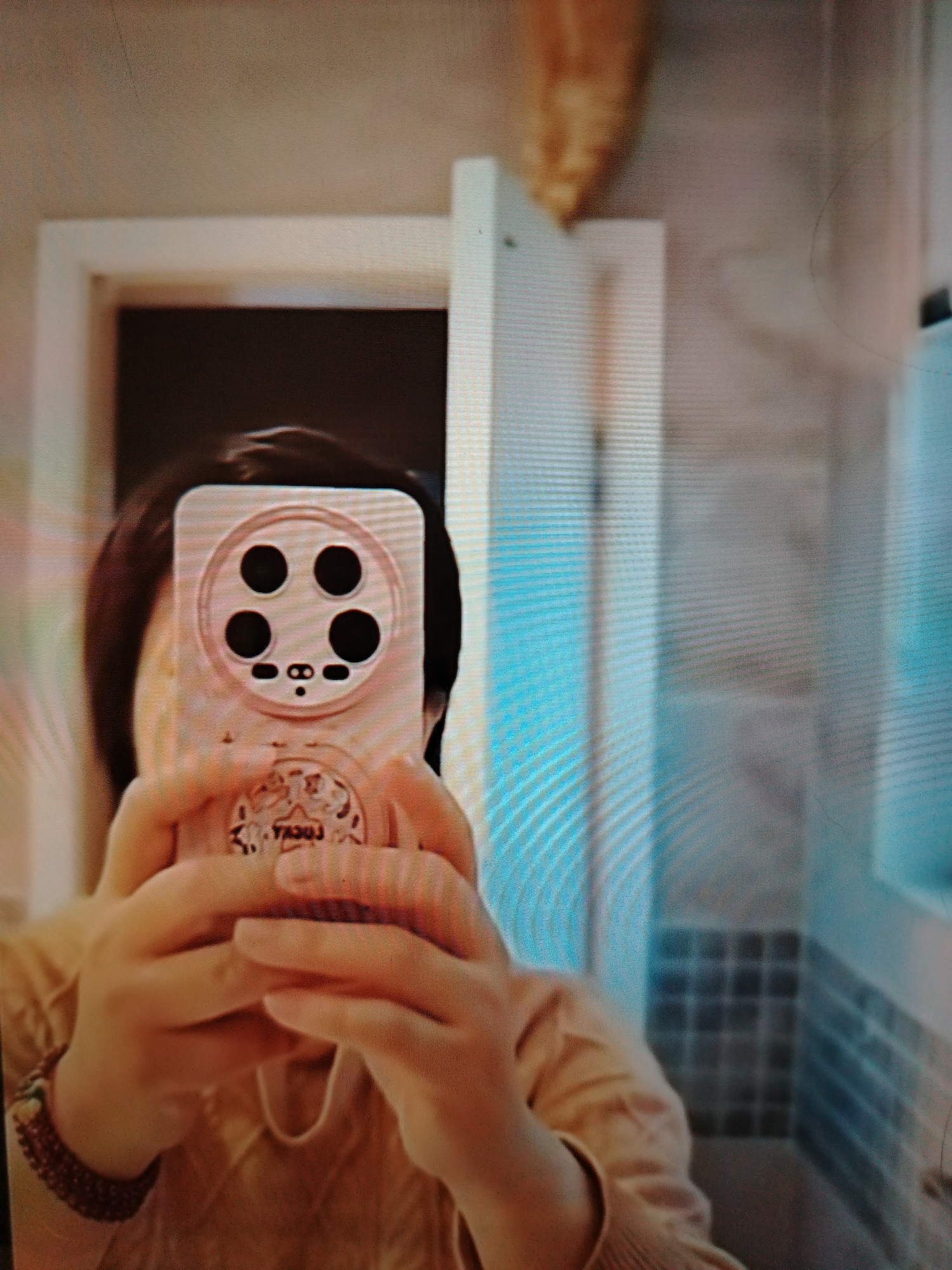 The image, captured in a mirror, shows a person holding a phone with a camera lens array that resembles a face. The background suggests an indoor setting, potentially a bathroom, with tiled walls visible. The lighting is diffused, and the overall image quality is low, likely due to the reflection and possibly the device used for capture. The scene is static, with no apparent interaction or dynamic action taking place. There is no information in the image that would indicate its location in Stavropol, Russia.