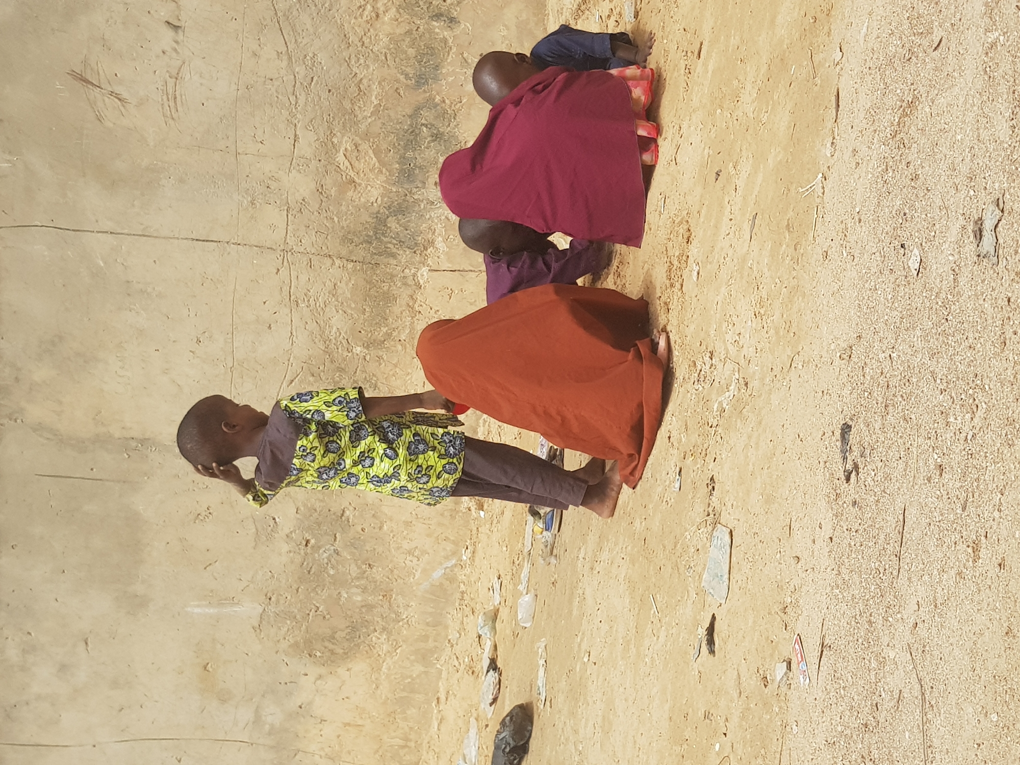 In Dutsinma B, Nigeria, the scene depicts a group of children on sandy ground near a wall. One child stands wearing a green and yellow patterned shirt, while three other children are kneeling, their heads bowed. The kneeling children are draped in maroon, purple, and orange cloth. There are various pieces of trash scattered across the sand.