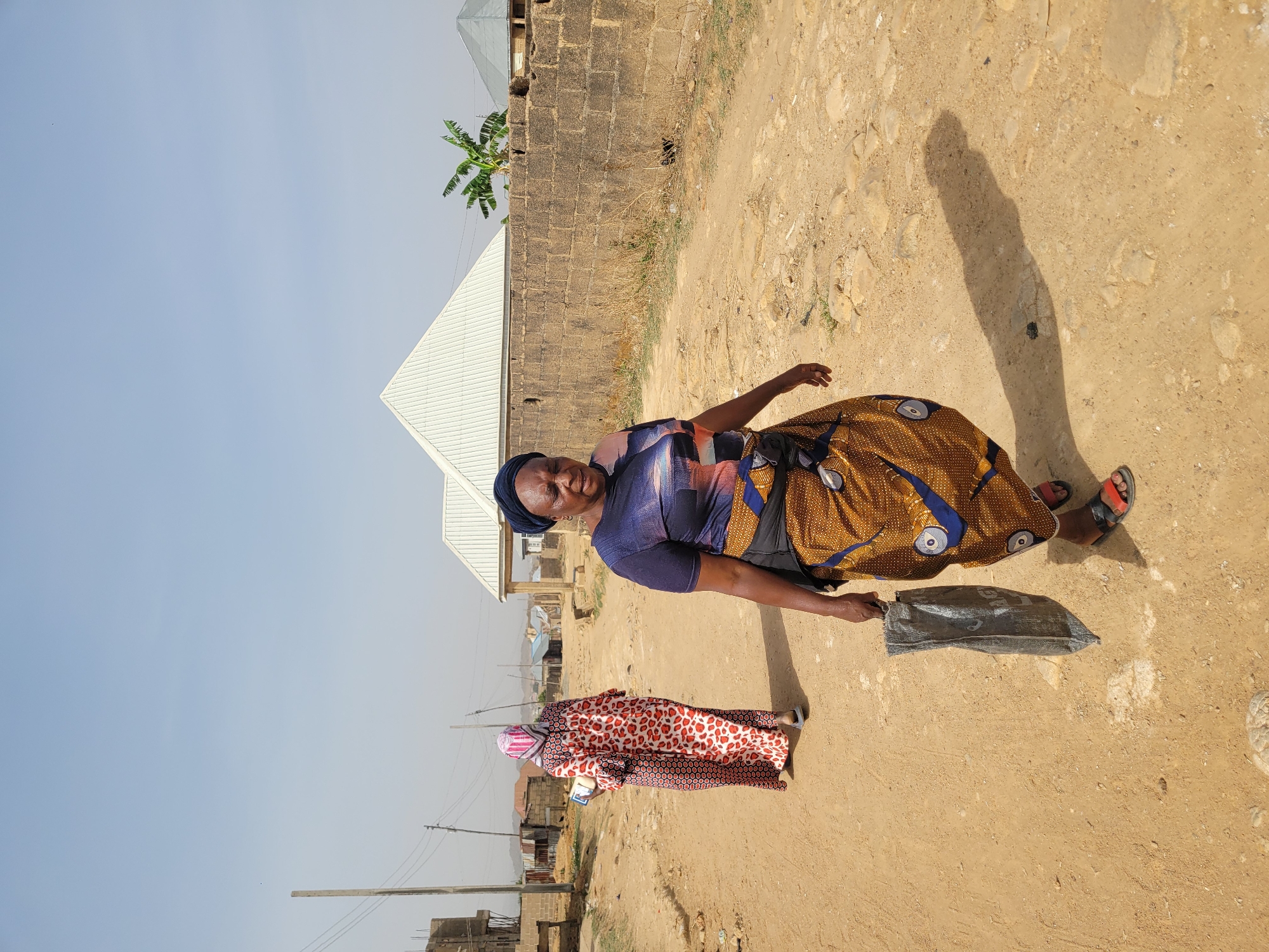 The image, taken in Jos, Nigeria, depicts two women on a sunlit, unpaved dirt path. The original image is oriented sideways (rotated 90 degrees counter-clockwise).
In the foreground, an older woman stands smiling towards the viewer. She is wearing a dark blue headwrap, a patterned short-sleeved top with blue, purple, and black tones, and a long wrapper skirt featuring a prominent brown, gold, and blue pattern with circular motifs. She wears red and black open-toed sandals and carries a dark, possibly black, bag in her left hand. Visible shadows on the path suggest a bright mid-morning or late afternoon sun.
Further down the path, another woman is walking away from the camera. She is dressed in a long, flowing garment with a distinctive red, black, and white leopard or animal print pattern, and a light-colored head covering with pink and white stripes. She appears to be holding a small, light-colored object in her left hand.
The scene is set outdoors in a residential area. To the left and behind the women, there are simple buildings constructed with concrete blocks and topped with light-colored corrugated metal roofs. A banana plant with large green leaves is visible behind the foreground woman. Utility poles and wires line the left side of the path, extending into the distance. The ground is dry, dusty, and uneven, consistent with an unpaved road or village pathway. The sky above is clear and light blue, indicating fair weather. No discernible text is visible on any objects or clothing.
