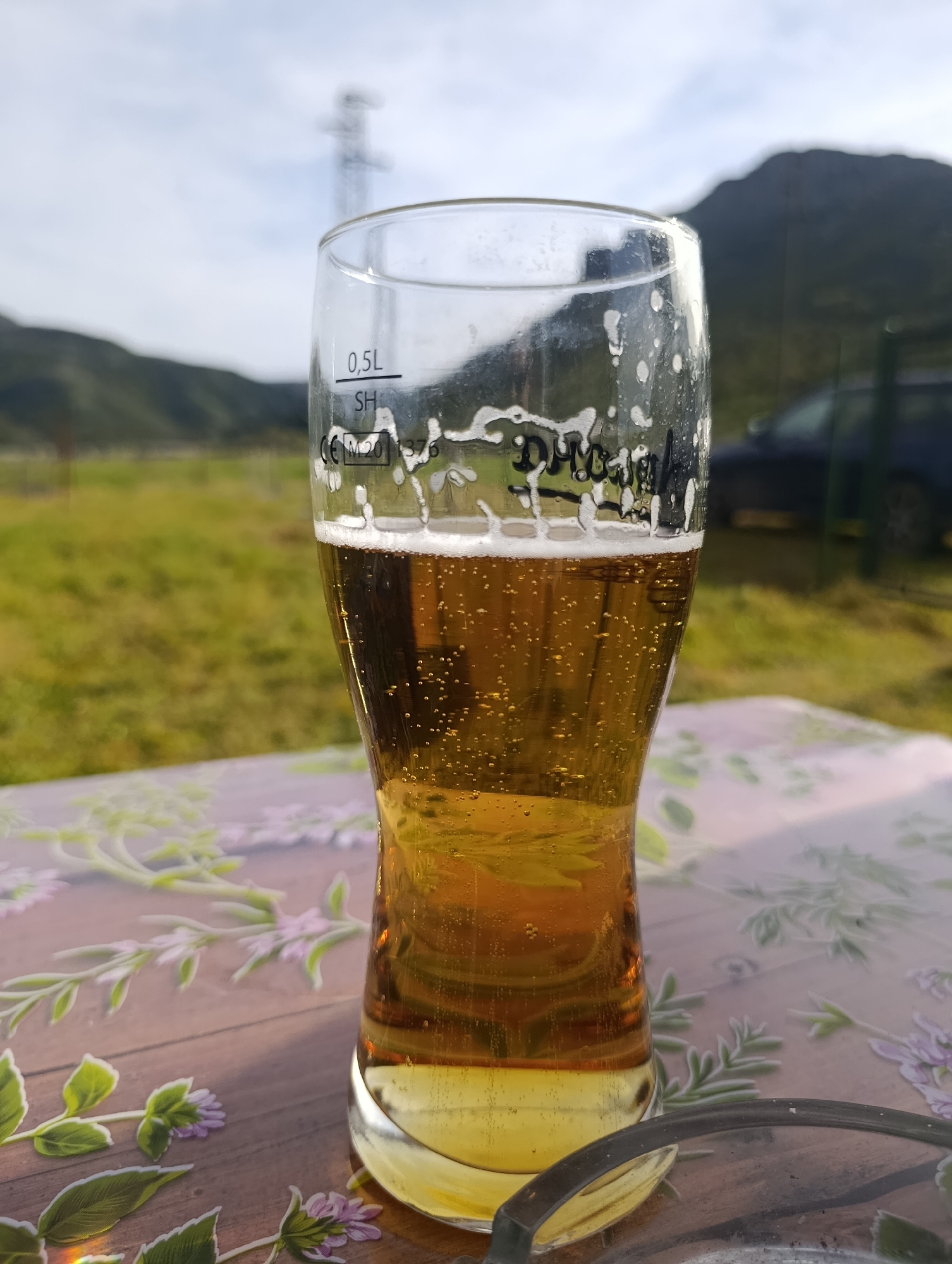 A half-filled beer glass sits on a table with a floral pattern in an outdoor setting. The beer is golden in color with visible bubbles and a frothy head. The glass itself has volume markings "0.5L" and "SH". In the background, there is a grassy field, mountains, a power line tower, and a dark-colored vehicle. The scene appears to be in the countryside, possibly near the city of Shiroko Pole, Bulgaria, as requested. There is an ashtray on the table in the foreground.