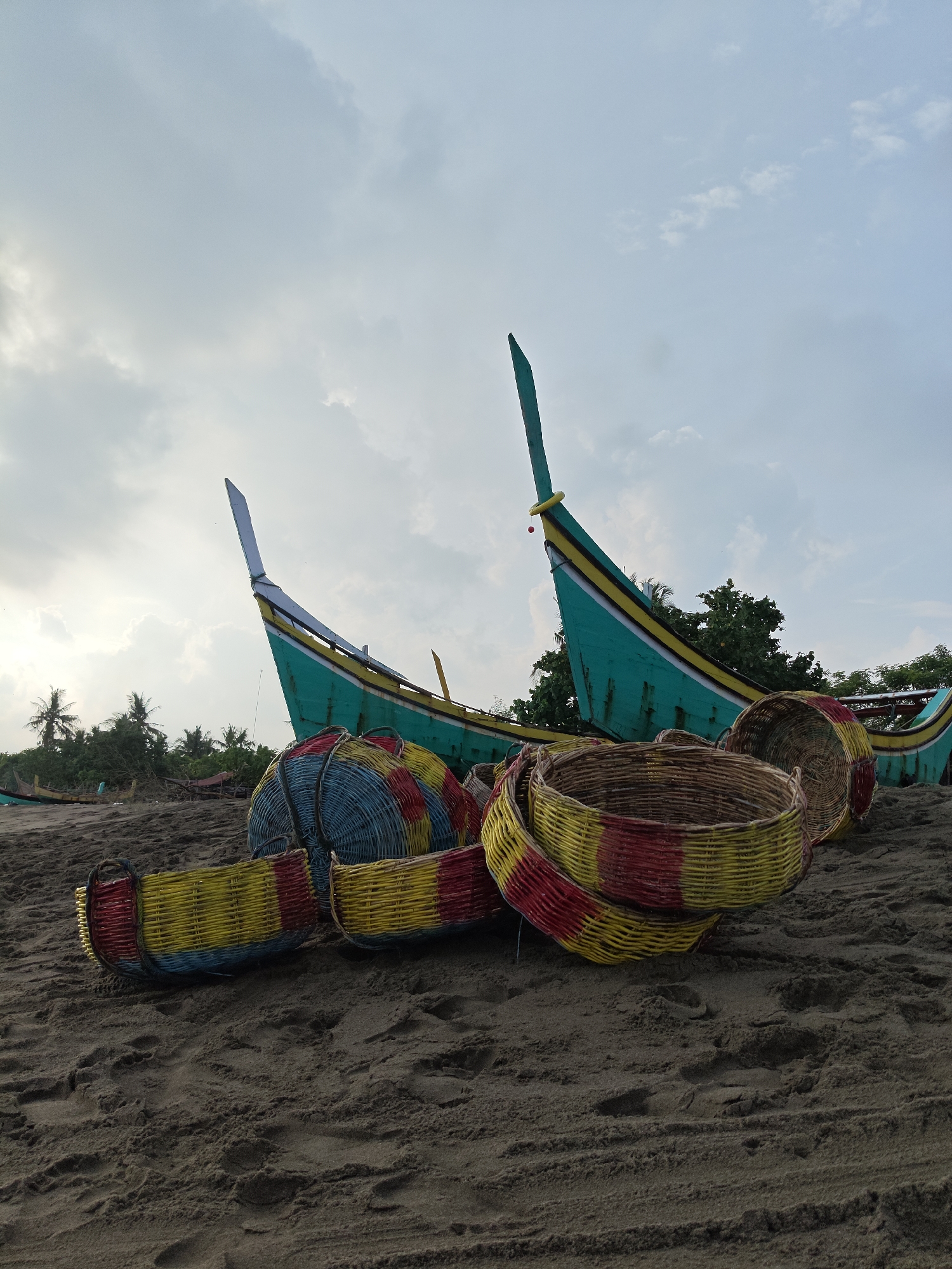 The image shows a beach scene in Curehtunong, Indonesia. In the foreground, there are several woven baskets of various sizes and colors, predominantly red, yellow, and blue. Two green and yellow fishing boats are positioned in the middle ground, with their bows pointing towards the sky. In the background, there is lush green vegetation, including palm trees. The sky is overcast with clouds. The scene appears to be a calm, coastal environment.