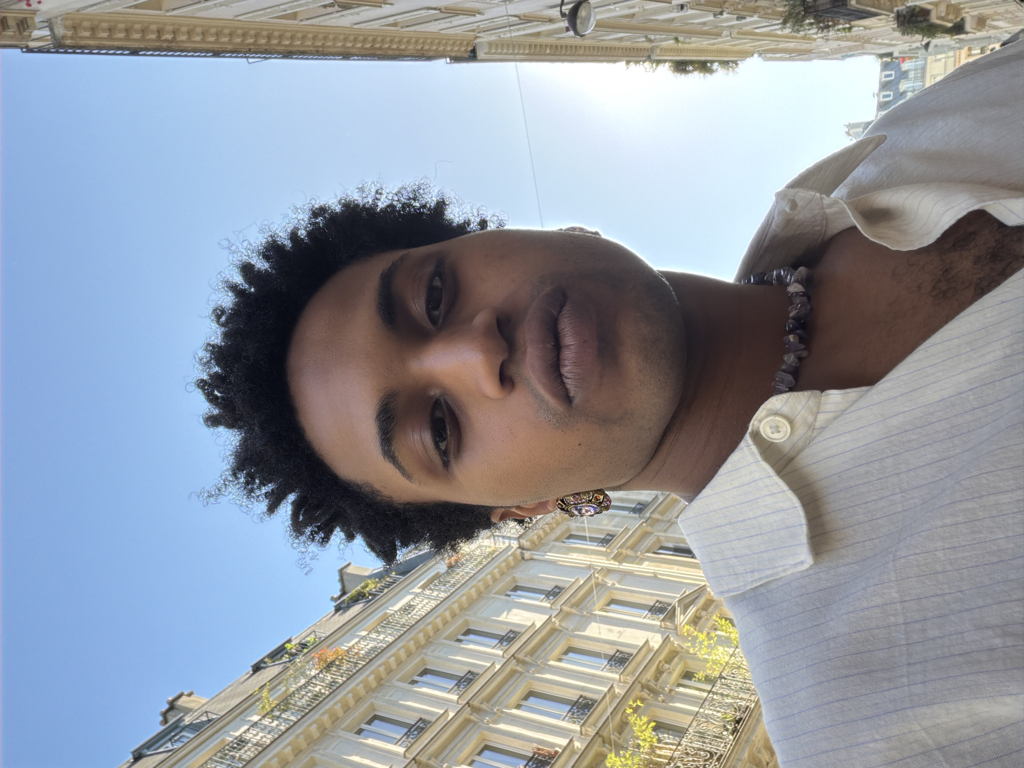 A low-angle portrait of a person with dark skin, curly hair, and facial hair stubble, framed against a Parisian backdrop. The subject is wearing a light-colored, button-up shirt and a beaded necklace and earrings. The background features classic Parisian buildings with symmetrical windows, balconies, and ornate architectural details. The sky is a clear, bright blue, indicating a sunny day in Paris.