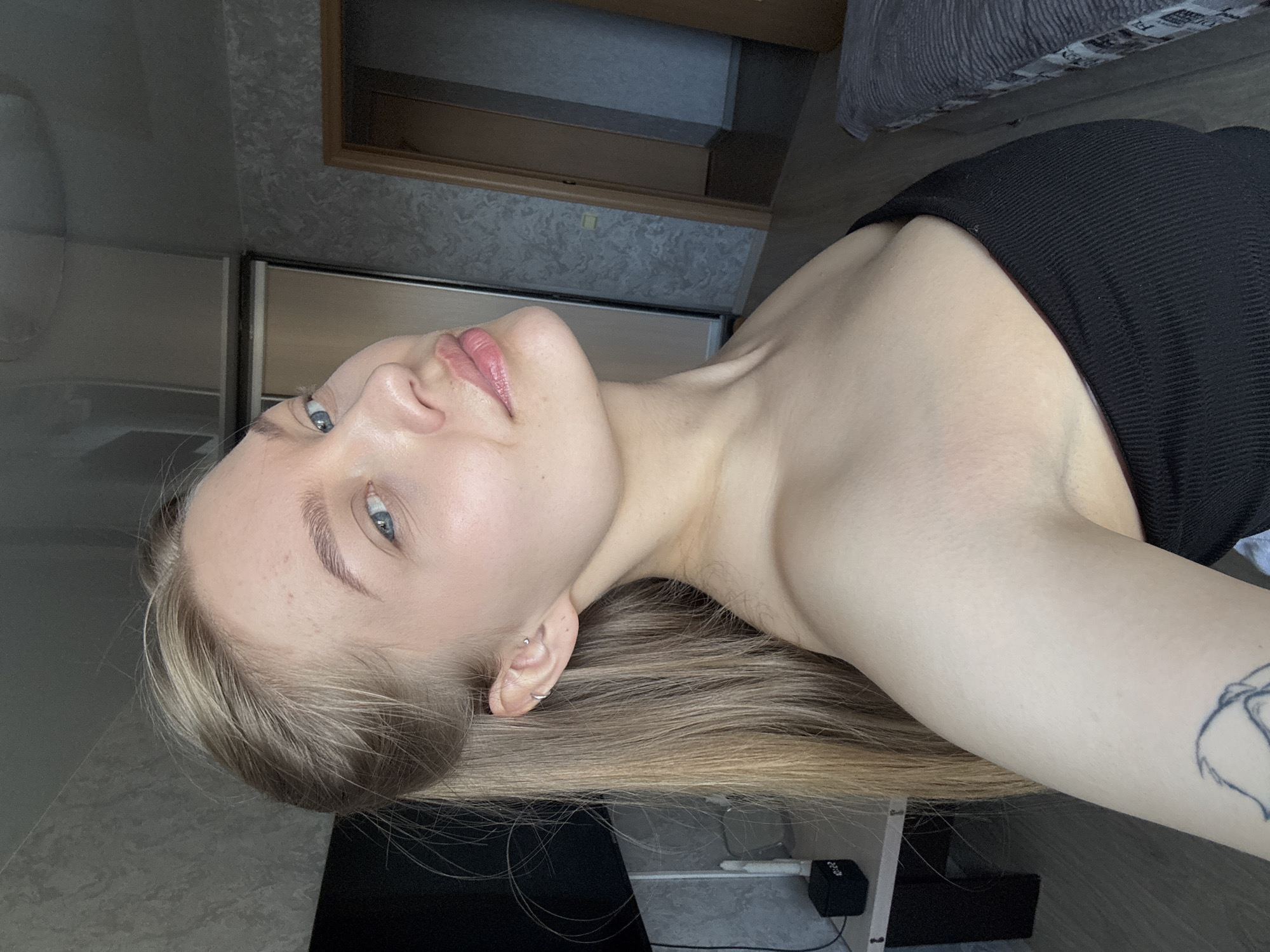 A young, fair-skinned woman with blonde hair lies on her side wearing a black tank top. She has a tattoo on her left arm. The background reveals a room with a wardrobe and a desk with computer accessories. The location is suggested to be Chaikovsky, Russia based on unspecified external information.