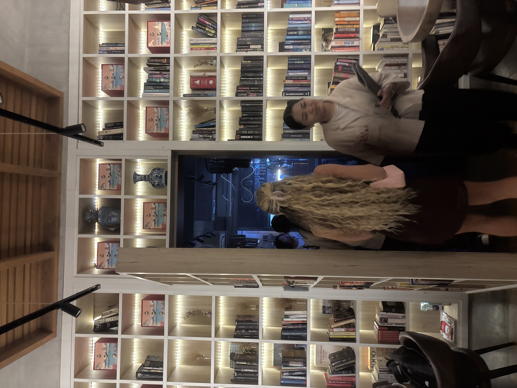 The image shows two individuals in what appears to be a bookstore or library with extensive shelving filled with books and decorative objects. The shelving is illuminated with embedded lighting. The individual closer to the camera has long, blonde, wavy hair and is wearing a brown dress or skirt. The other individual, facing the camera, is wearing a light-colored shirt and dark pants, holding a tablet or book. The shelves behind them are arranged in a grid-like pattern, filled with books of various sizes and colors, as well as decorative boxes and vases. The location appears to be a well-lit, indoor space. OCR identified various book titles, some in English and some Cyrillic, including "WILL", "THE ARTZRITZ", "AIRSIDE", and "SONIA". I could not definitively determine the location, but information on similar-appearing libraries in North Kuta, Indonesia was found.