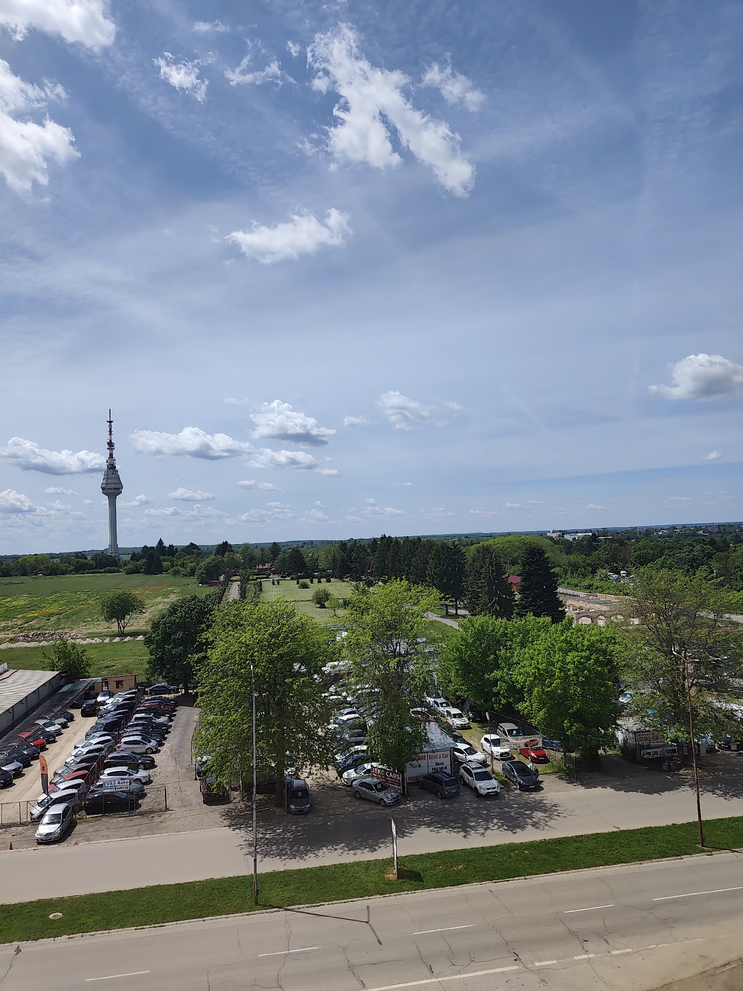 The image presents a high-angle view of a landscape in Dobrich, Bulgaria. In the distance, a prominent telecommunications tower dominates the skyline. A large open field stretches out in front of the tower, bordered by trees and green vegetation. Closer to the viewer are several parking lots filled with various cars. A road runs along the bottom of the frame, showing cracks in the asphalt. The sky is partially cloudy with patches of blue. The lighting is bright, suggesting a sunny day.