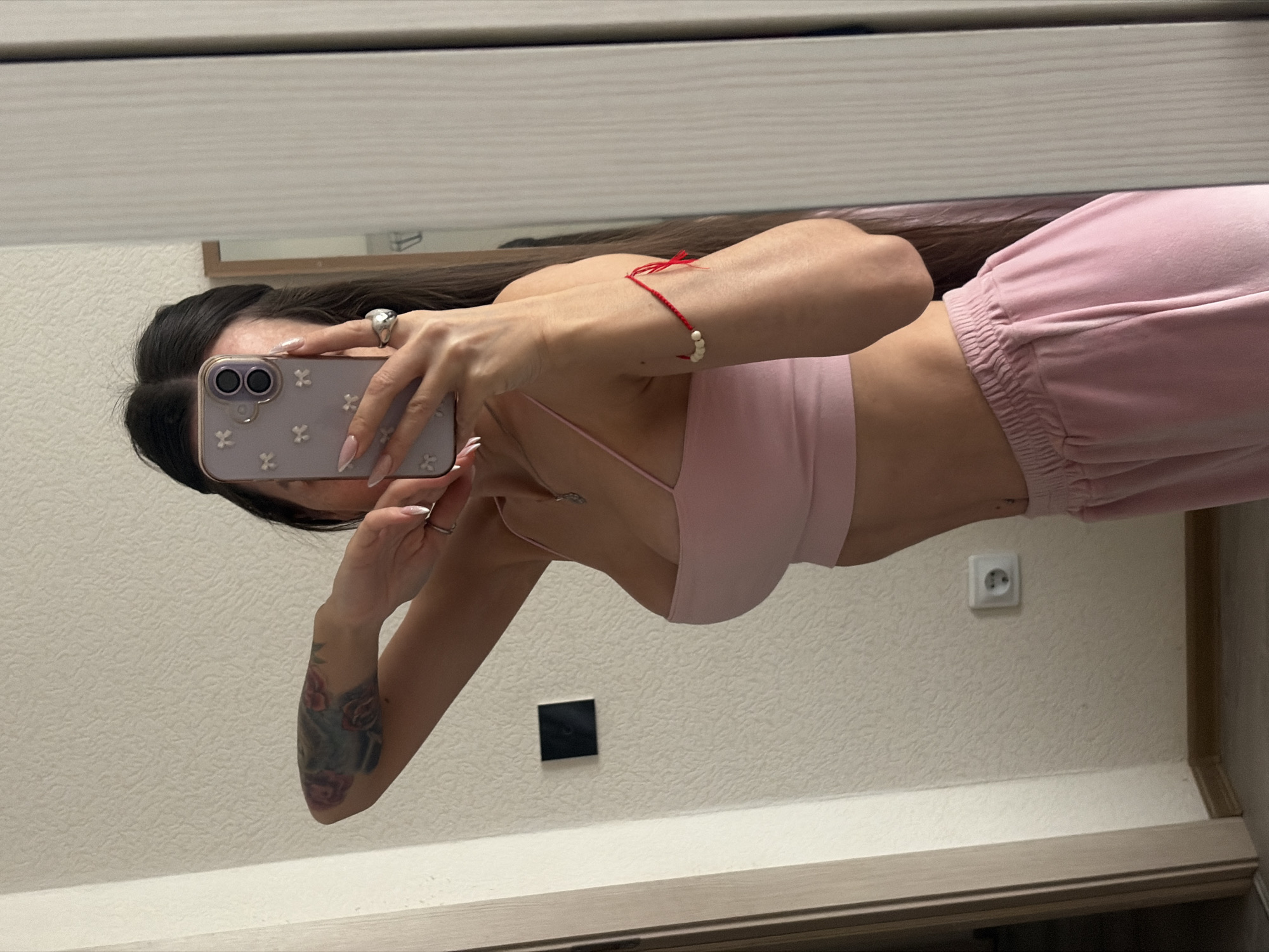 A woman is taking a mirror selfie. She is wearing a pink top and pants. She has a tattoo on her left arm and a bracelet on her right wrist. She is holding a cell phone in her right hand. The background is a wall with an electrical outlet and a light switch. The location is not specified.