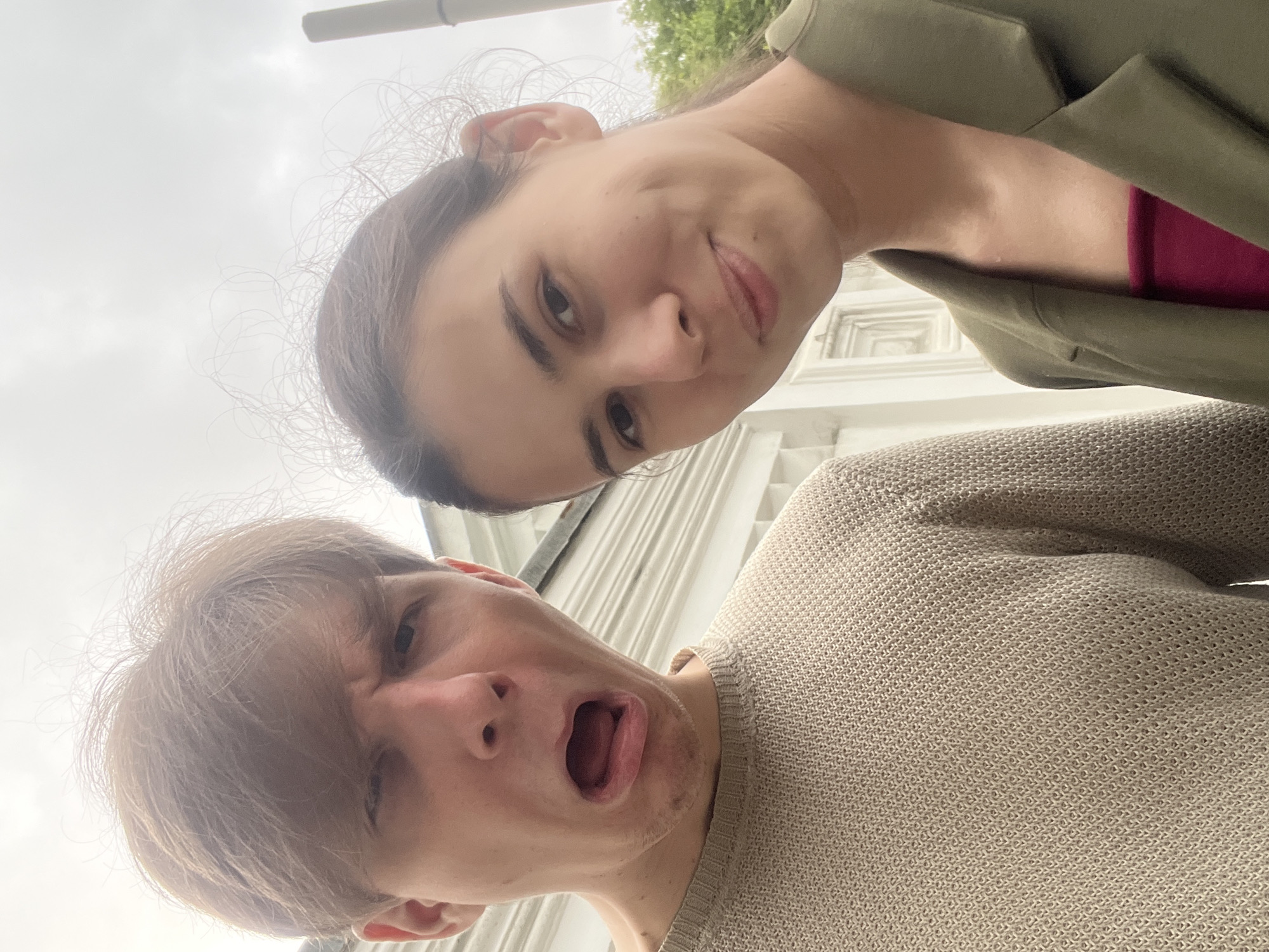 A selfie is taken in an outdoor environment, with a light-colored building and a cloudy sky in the background. A young man and woman are the subjects. The man, on the left, is making a surprised or disgusted face, with his mouth wide open and eyes squinted. He is wearing a light-colored, knitted sweater. The woman, on the right, is smiling gently, looking towards the camera. She is wearing a green jacket over a dark pink top. The building behind them appears to be a white, multi-story structure with architectural details. A vertical, grey object, possibly a chimney or pole, is visible in the upper right corner of the image. Green foliage from trees can be seen in the background on the far right.