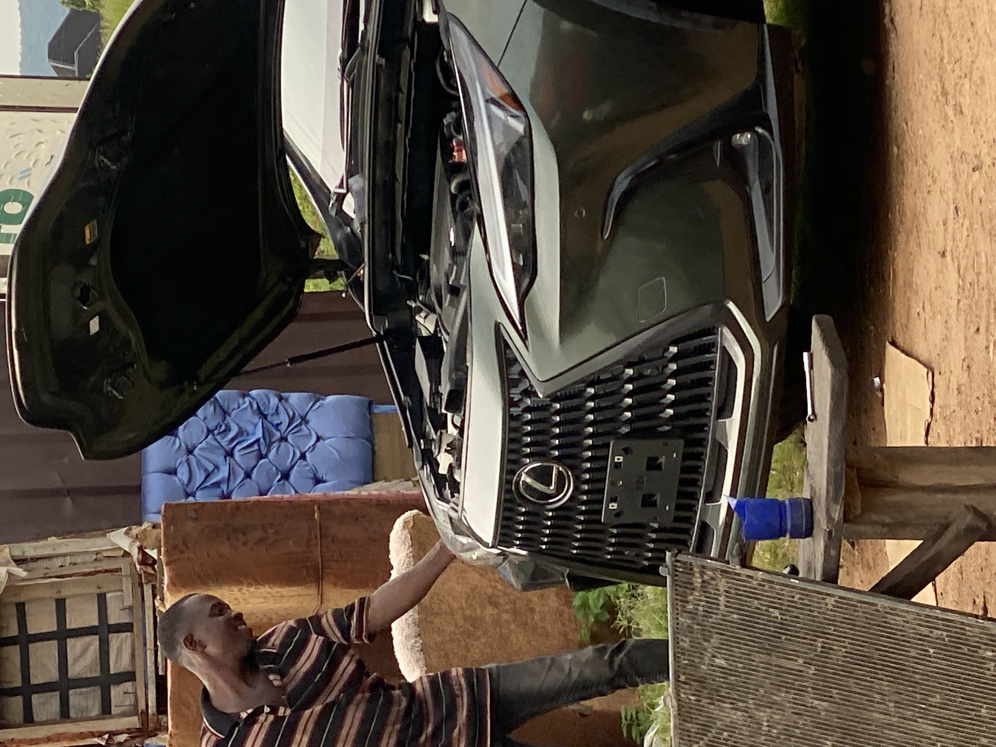 A man in a striped shirt and jeans is seen next to a dark green Lexus SUV with its hood open, indicating he is working on the engine. The car is parked outdoors on a dirt surface, with a wooden table and a piece of equipment that looks like a radiator to the left. In the background, there are stacked items that appear to be mattresses or cushions and a portion of a shipping container. The scene is likely in Jalingo, Nigeria, given the context of the question, though the image itself does not provide specific geographical markers to confirm the location. The lighting suggests it is daytime.
