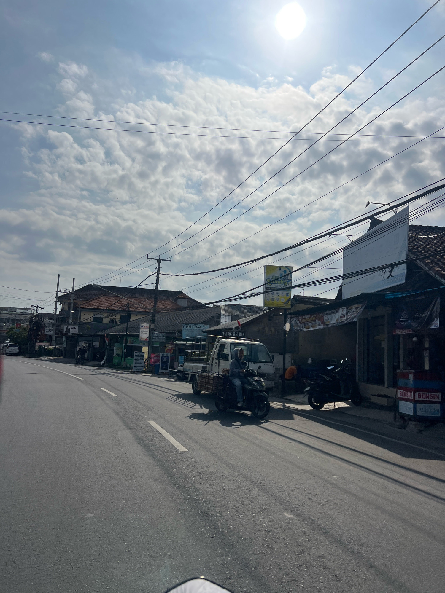 The image depicts a street scene in Denpasar, Indonesia, under a bright, cloudy sky. The road, paved with asphalt and marked with white lines, curves towards the left. On the right side of the road, a row of businesses with awnings lines the street. These businesses appear to be shops or small establishments, with some displaying signage. One sign reads "BENSIN," indicating a fuel station.
Several vehicles are present. A white pickup truck with a wooden flatbed is parked on the right side of the road. In front of it, a person is riding a dark-colored scooter, heading towards the foreground. Another dark-colored scooter is parked behind the pickup truck. Further down the street, a white car is visible in the distance on the left.
The environment is characterized by overhead power lines crisscrossing the sky, supported by utility poles. The buildings are modest in construction, with tiled roofs. The overall atmosphere suggests a typical urban street in Indonesia on a sunny, yet partly cloudy day.