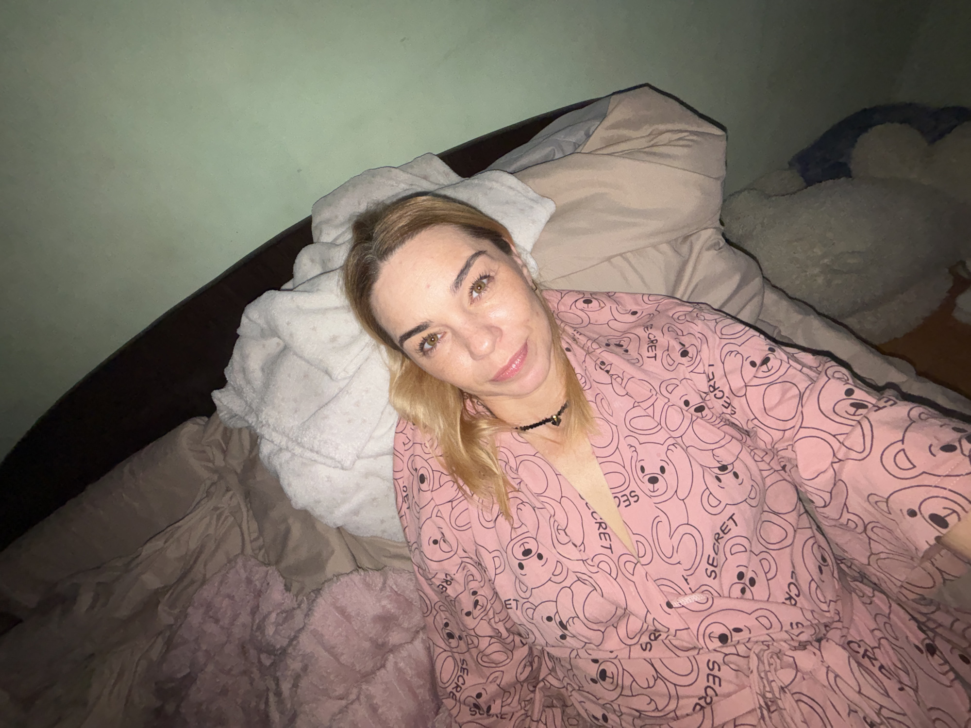 A light-skinned woman with blonde hair, wearing a pink robe with a teddy bear pattern, lies on a bed. She is wearing a black beaded necklace. The bed is covered with various blankets and pillows in shades of pink, beige, and white. A portion of a dark wooden headboard is visible. The background is a wall painted a light green color. The scene appears to be indoors, in a bedroom.